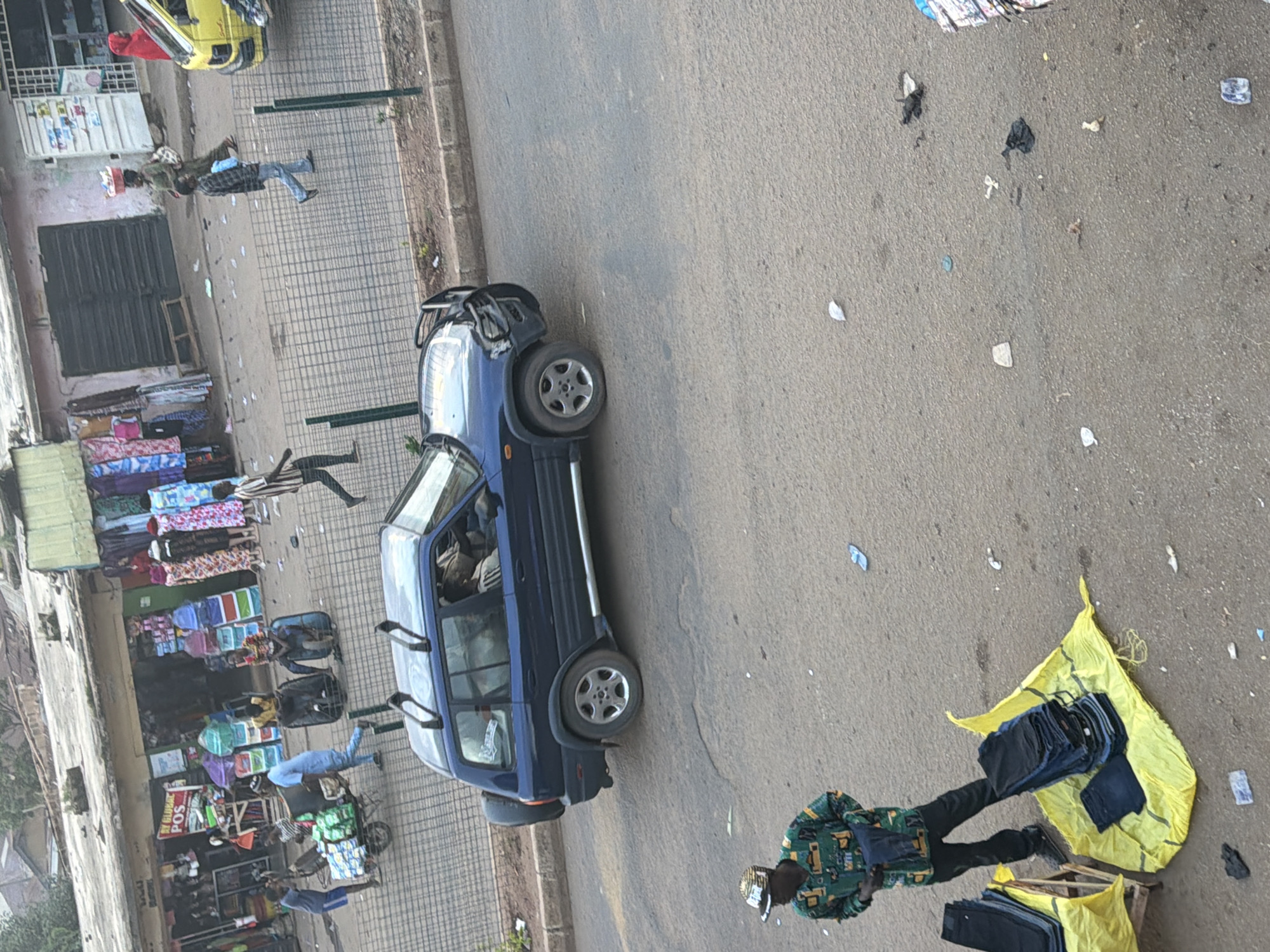 The image depicts a street scene in Bukuru, Nigeria. A blue van is parked on the right side of the road, with several people visible inside and around it. A man wearing a green patterned shirt and a white and black hat is standing near the van, looking towards the right. In the foreground, a yellow cloth is spread on the ground, displaying stacks of folded dark-colored trousers, presumably for sale. Another man in a similar green patterned shirt is visible on the left, partially obscured by the van.
To the left of the van, a market or shop area is visible, with various goods displayed, including stacks of colorful fabrics and what appear to be boxes or containers. A yellow vehicle is parked further back on the left. People are seen interacting within this market area, with some individuals standing and others seemingly engaged in activities related to the shops. The ground is paved and appears to be a street or sidewalk, with some debris scattered around. The scene suggests a bustling commercial area, likely during daylight hours, though the specific time of day and weather conditions are not definitively ascertainable from the image. No specific text is clearly legible in the image.