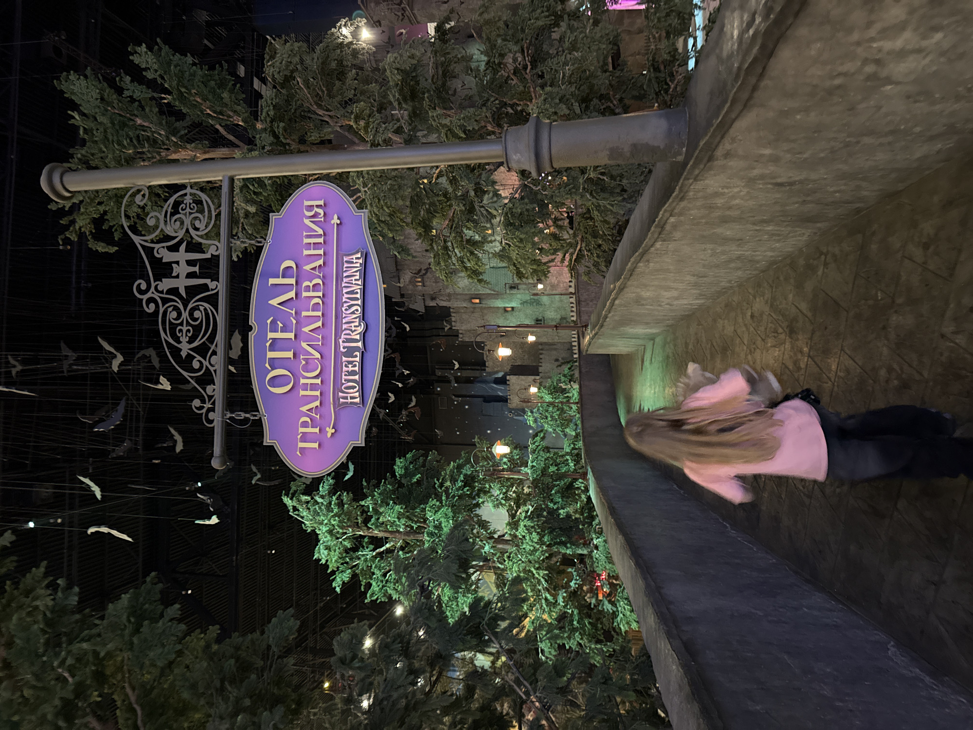 The image captures a dimly lit, themed indoor environment, likely an attraction related to "Hotel Transylvania" in Moscow, Russia. A person, possibly a child or young individual with long blonde hair, wearing a pink top and dark pants, is visible on the right, walking along a concrete pathway or bridge. Their figure is blurred, indicating motion.
On the left, a prominent purple, oval-shaped sign hangs from an ornate grey metal bracket and pole. The sign displays text in two languages: "ОТЕЛЬ ТРАНСИЛЬВАНИЯ" (OTEL' TRANSIL'VANIYA), which translates to "Hotel Transylvania," is written in large Cyrillic letters, with "HOTEL TRANSYLVANIA" in smaller English letters below.
The scene is heavily decorated with artificial green trees and foliage, creating a forest-like atmosphere. Numerous small, dark cut-out shapes resembling bats are suspended in the dark background area near the sign, contributing to the themed ambiance. Scattered throughout the environment are decorative lights and lamps, casting warm glows against the dark setting. The overall impression is that of a fantastical, immersive experience, likely designed to evoke a spooky yet playful mood.