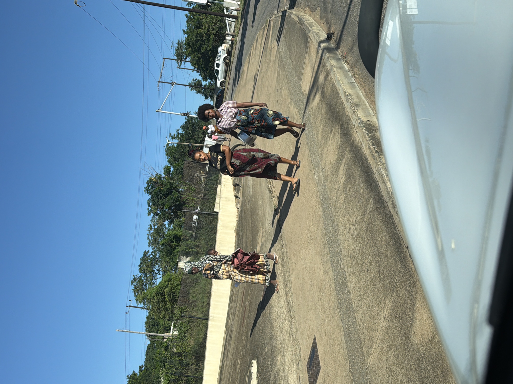 The image captures an outdoor scene on a bright, sunny day, likely taken from inside a vehicle. Three women are walking along a paved surface that appears to be a roadside or a wide curb.
In the foreground, a woman is seen from the waist up, wearing a dark t-shirt with the word "Betway" visible on it, and a patterned skirt. She is looking towards the camera. Behind her, another woman is walking, dressed in a plaid shirt and a patterned skirt, carrying a large, metallic bowl in her hand. She is also looking in the general direction of the camera. Further back, a third woman, wearing a patterned dress or skirt and a head covering, walks with a baby securely wrapped on her back. All three women appear to be wearing sandals or are barefoot.
The ground is a grey, textured asphalt or concrete, with clear shadows cast by the people, indicating strong sunlight. To the far right, the white hood and windshield edge of the vehicle from which the photo was taken are visible. In the background, there are green trees, utility poles with numerous power lines stretching across a clear blue sky, and a white pickup truck parked further down the road. The overall setting suggests a semi-urban or rural area. There are no strong emotional expressions discernible on the women's faces; they appear to be going about their daily activities.