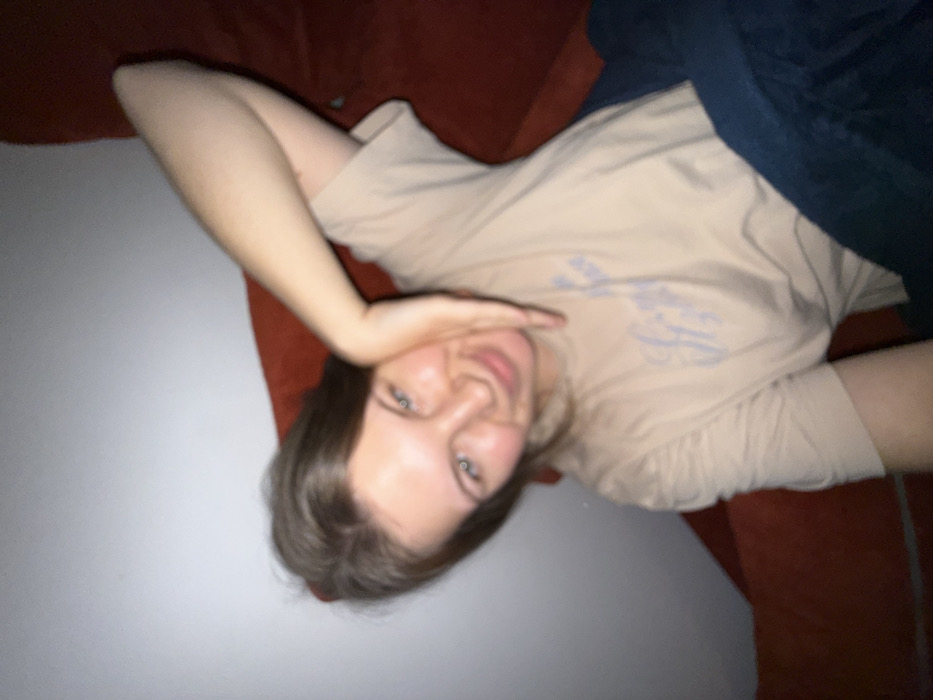 A young woman with light skin and gray eyes lies sideways on a dark orange sofa. She wears a tan t-shirt with a faint design and blue jeans or a blanket covers part of her torso. Her head rests on a portion of the sofa, and one hand is positioned near her face. The background includes a plain white wall.