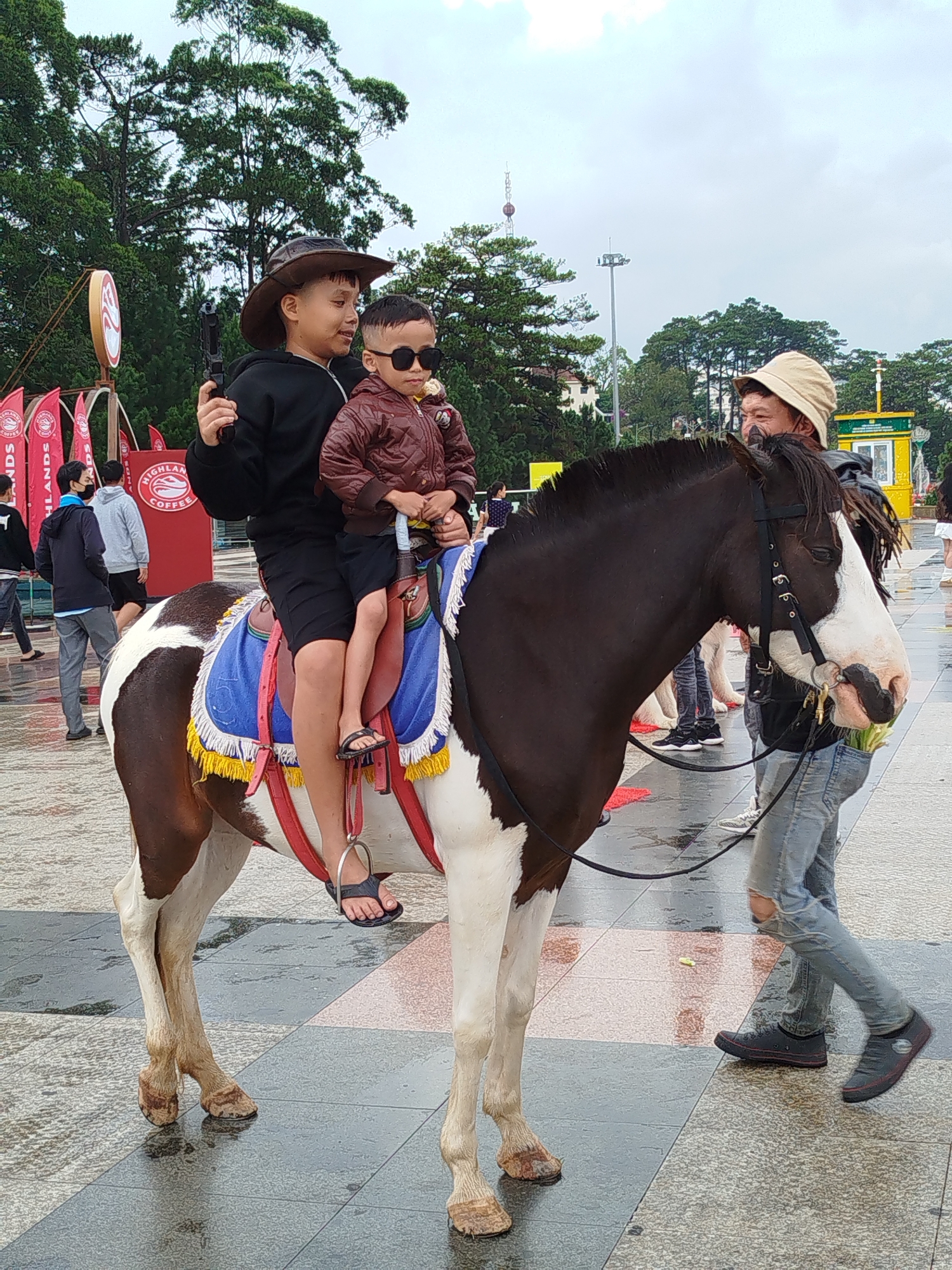 The image depicts two children riding a brown and white horse in what appears to be a public square. The older child, wearing a brown cowboy hat and black clothing, holds what looks like a toy gun. The younger child, wearing sunglasses and a brown jacket, sits in front. The horse has a blue saddle and yellow fringe. A man in a light-colored hat and jeans holds the horse's reins. The background suggests an urban environment, with trees, buildings, and people walking in the distance. Signage with the words "HIGHLANDS COFFEE" suggests the location is Da Lat, Vietnam, where this coffee chain is prevalent. The ground appears wet.