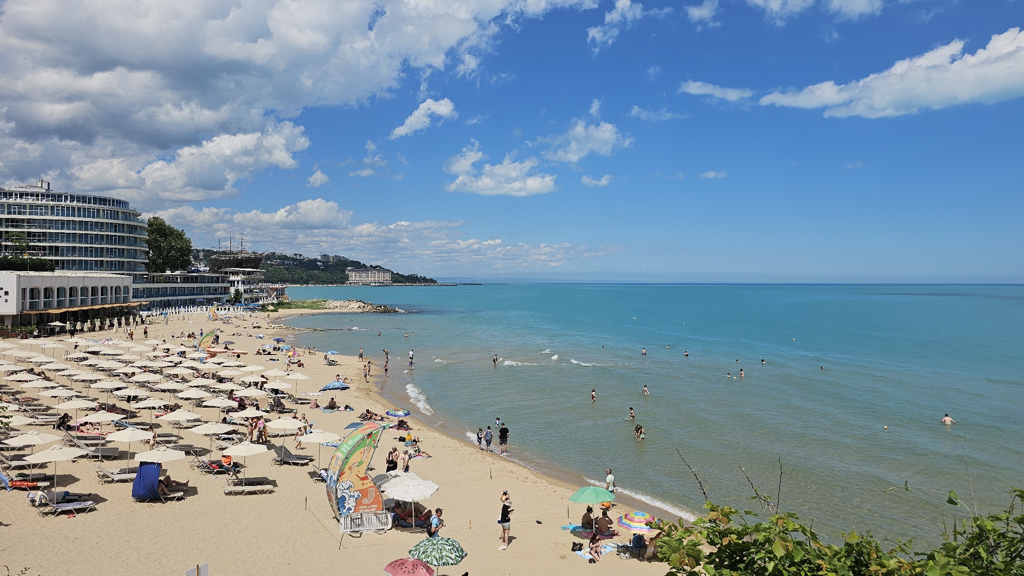 The image depicts a sun-drenched beach on the Black Sea coast, likely in the resort area of Varna, Bulgaria. A large, curved, multi-story hotel complex is visible on the left, with its white facade and numerous balconies. In the distance, along the coastline, other buildings and a large sailing ship are discernible.
The beach itself is covered with a multitude of tightly packed, cream-colored umbrellas and lounge chairs, creating a dense pattern. Several people are scattered across the sand, some lounging under the umbrellas, others walking, and a few are near the water's edge.
The sea is a vibrant turquoise, with gentle waves lapping at the sandy shore. Numerous individuals are in the water, swimming or wading. The sky is a bright blue, punctuated by fluffy white clouds.
In the foreground, on the right side of the image, there is dense green foliage, suggesting the viewpoint is from a slightly elevated position overlooking the beach and sea. Colorful beach umbrellas, including a large green one and smaller patterned ones, are visible closer to the water, indicating a more relaxed and perhaps less organized section of the beach.