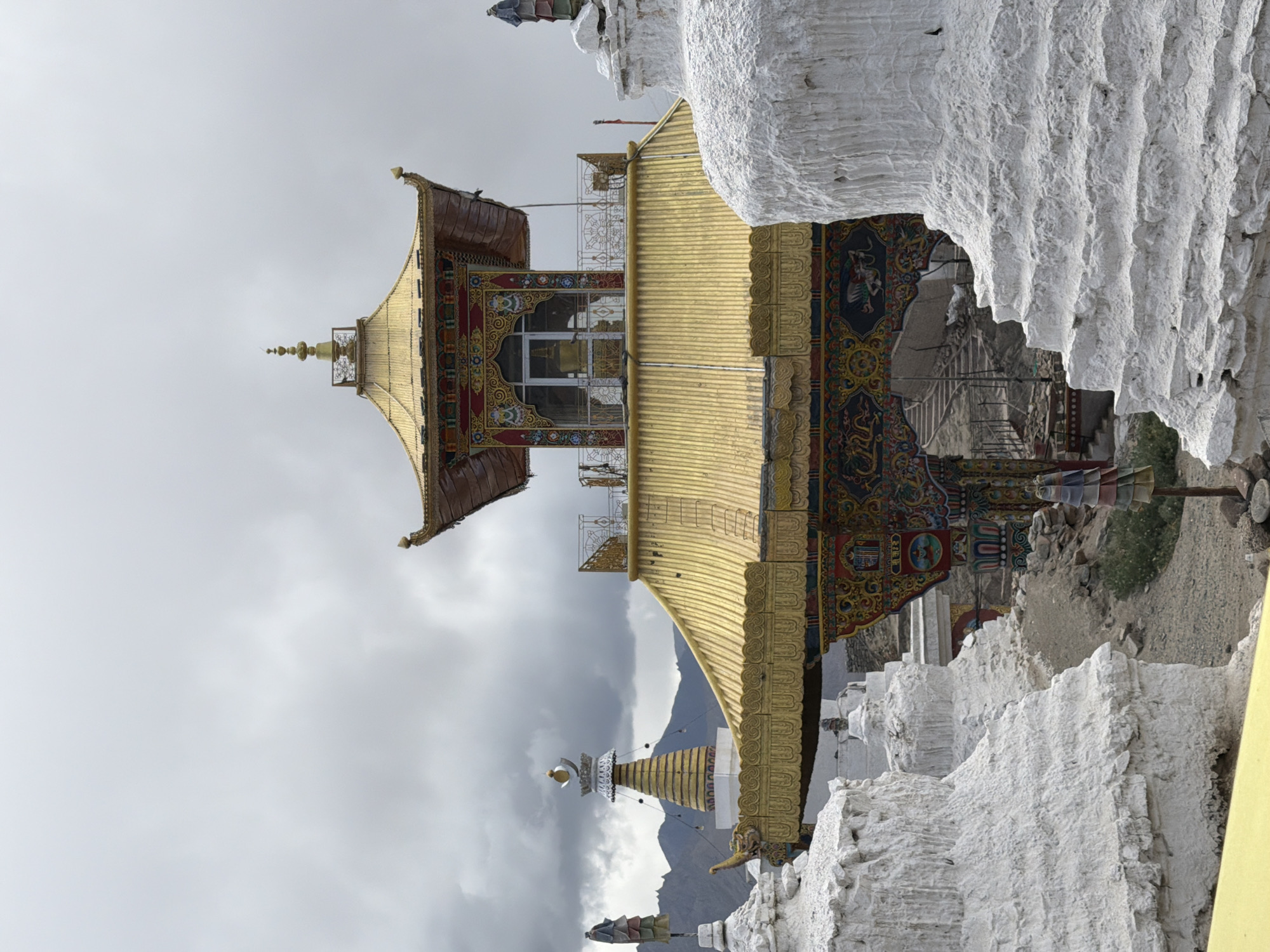 The image depicts a Buddhist monastery structure, likely a stupa, in Leh, India. The foreground is dominated by several whitewashed, tiered structures, which are characteristic of stupas. These structures have a rough, textured surface and are partially obscuring the central building.
The central architectural element is a multi-tiered building with a golden, ornate roof. The roof is intricately designed with what appears to be golden shingles or tiles, and it has several decorative finials. Below the golden roof, there is a section with colorful, detailed murals or paintings depicting various motifs, possibly including dragons and other traditional symbols. This section also features decorative pillars and architectural embellishments.
Above the main golden roof, a smaller, pagoda-like structure is visible, also topped with a golden spire and finial. Windows can be seen on this upper level. The entire structure is set against a backdrop of a cloudy, overcast sky, with distant mountains faintly visible. There are also several smaller white stupa-like structures and decorative elements scattered around the site, including what looks like prayer flags. The ground appears to be a mixture of gravel and dirt. The scene suggests a religious or cultural site, given the architectural style and the presence of stupas.
