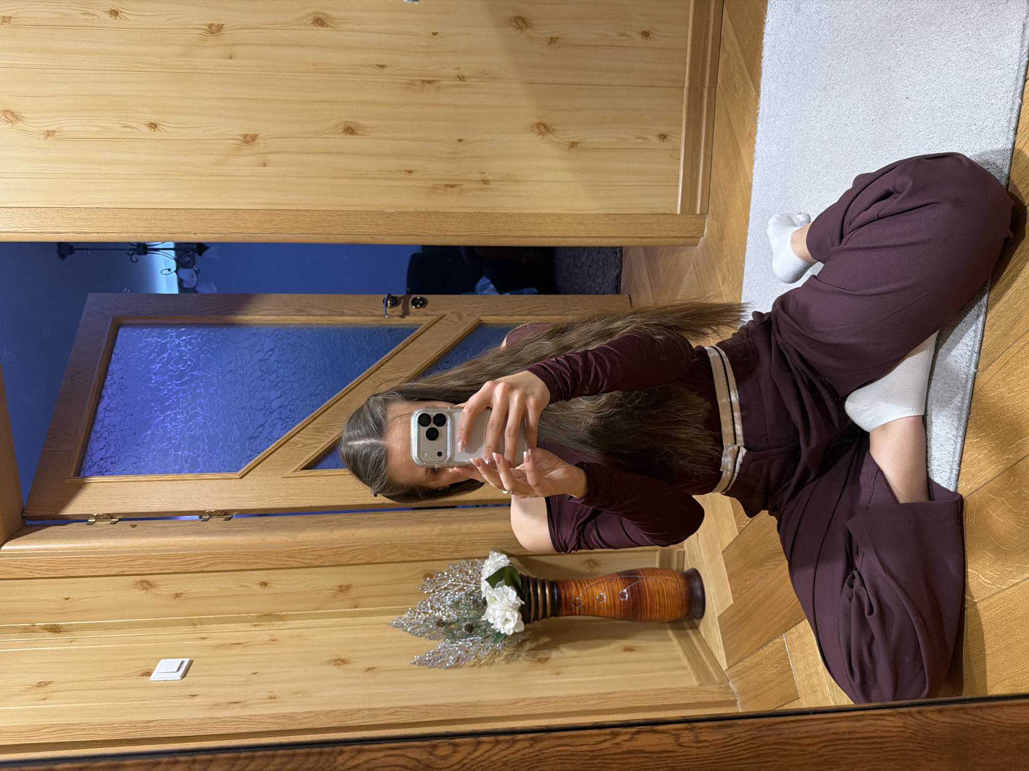 A person kneels on a gray rug, clad in dark purple clothing and white socks, taking a mirror selfie with a white smartphone. The reflection shows a wooden door behind them, leading to a dimly lit room. A wooden vase containing white flowers and silver decorative branches sits in the foreground. The location may be Buhovo, Bulgaria, as specified in the query. The surface on which the person kneels is wooden parquet flooring.