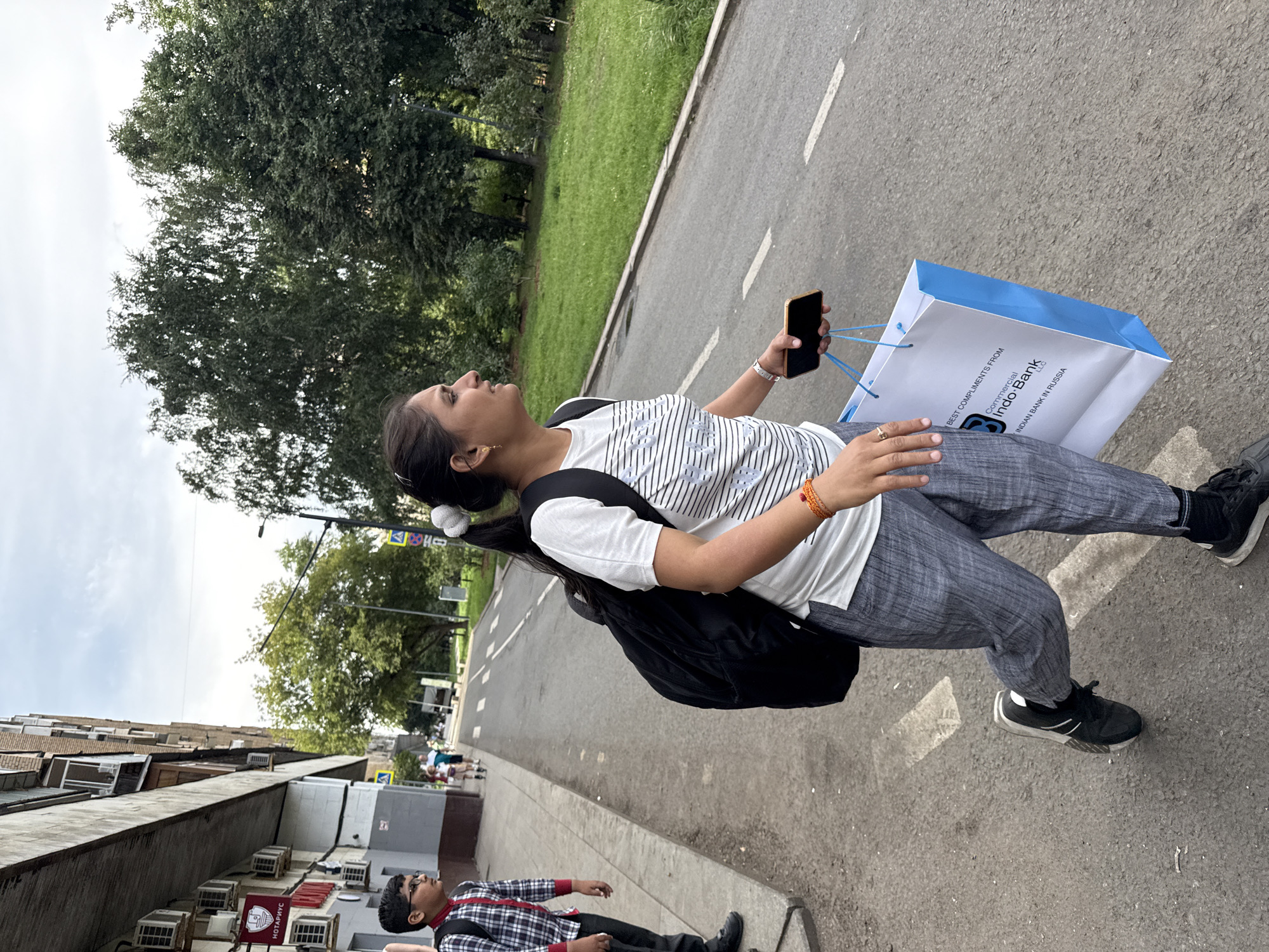 A woman with her hair in a ponytail is walking on a sidewalk next to a road. She is wearing a white and black striped t-shirt, gray checkered pants, black sneakers, and a black backpack. She is holding a smartphone in her right hand and a white shopping bag with blue handles in her left hand. The bag has the logo of "Commercial Indo-Bank LLC" and "INDIAN BANK IN RUSSIA" printed on it. A young boy wearing a red and black checkered shirt, black pants, and a backpack is standing on the sidewalk to the left of the woman. He is looking towards the road. The scene takes place on a city street in Moscow, Russia, with a building on the left and trees and a grassy area on the right. The sky is cloudy.