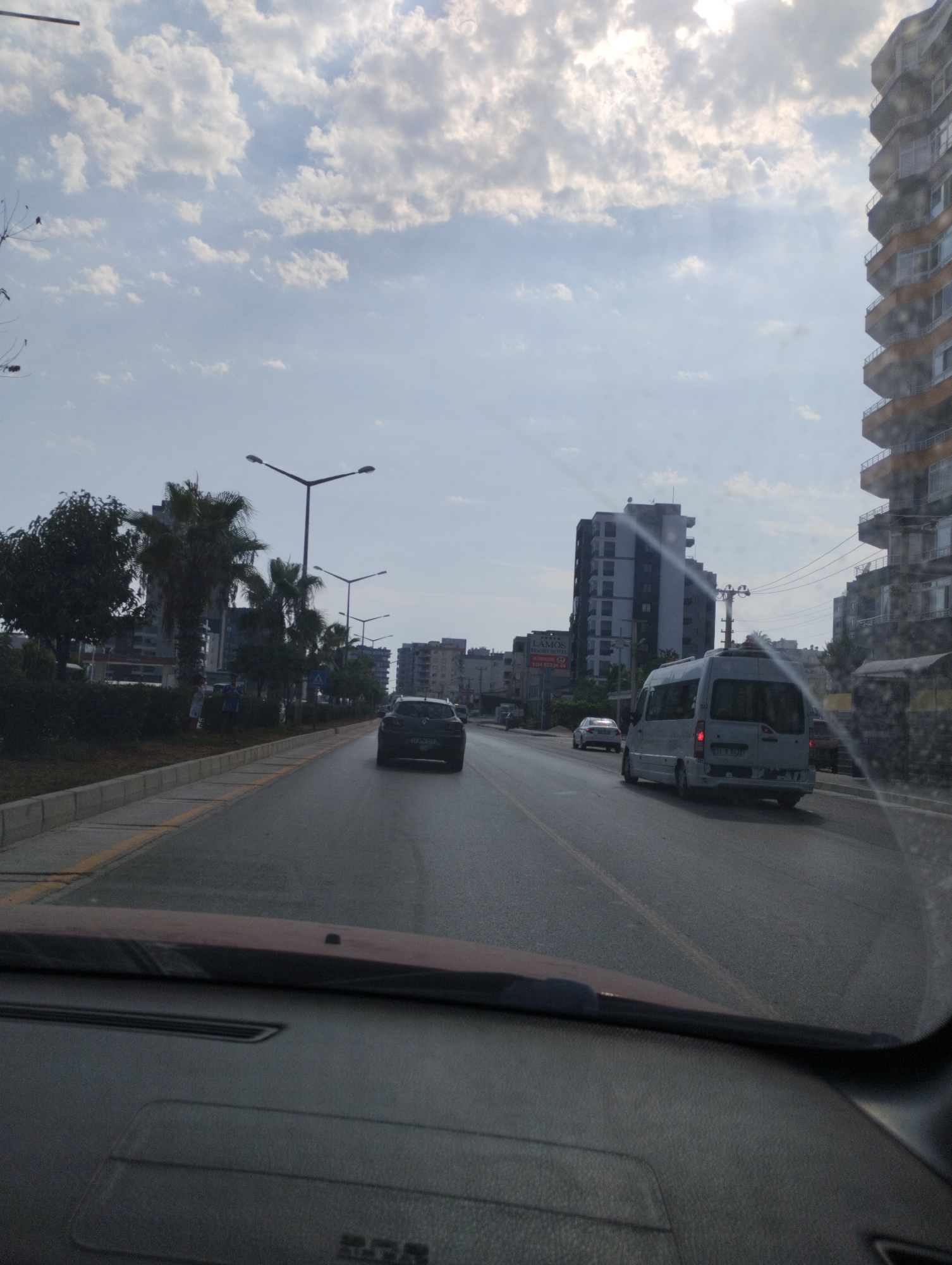The image captures a view from inside a vehicle, looking forward through the windshield at a city street in Mezitli, Turkey. The sky above is partly cloudy with bright sunlight filtering through. On the right side of the road, a tall, modern apartment building with balconies is prominently visible. The street itself is paved and appears to be a multi-lane road. Several vehicles are on the road, including a white van in the middle lane, a dark-colored car ahead of it in the right lane, and another car in the distance. On the left side of the road, there are trees, including palm trees, and streetlights. Buildings and more trees are visible further down the street. The foreground shows the dashboard of the vehicle from which the photo is taken, with a portion of the car's interior visible.