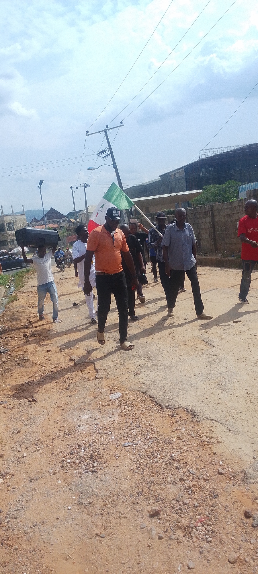 The image shows a group of people walking along a dirt road in what appears to be a city environment. The location is potentially Lokoja, Nigeria, based on the presence of individuals who could be described as African, along with a flag bearing the word "KADDO". The road is unpaved and appears to be in disrepair. The group includes men dressed in casual clothing. One man is carrying a large black box over his head. Another man in an orange shirt is at the front. A man in a red shirt is on the right. There is a flag with green, white, and red colors being carried in the middle of the group. In the background, there are buildings, power lines, and parked vehicles, indicating an urban or semi-urban setting. The sky is cloudy.