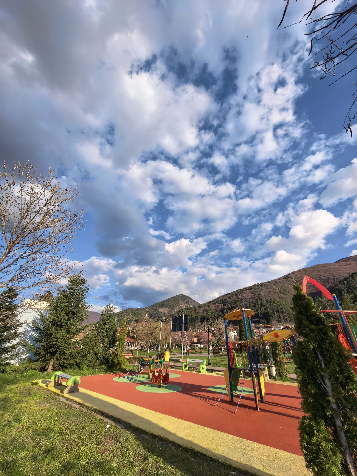 The image shows a playground in a park located in Unknown, Unknown. The playground features colorful equipment including slides, climbing structures, and swings on a red and yellow rubber surface. Green benches are located near the playground equipment. The park has green grass and various trees, including evergreen trees and a bare-branched tree. A mountain range is visible in the background. The sky is blue with scattered white clouds and some darker storm clouds. A utility pole is also present. People can be seen in the background.