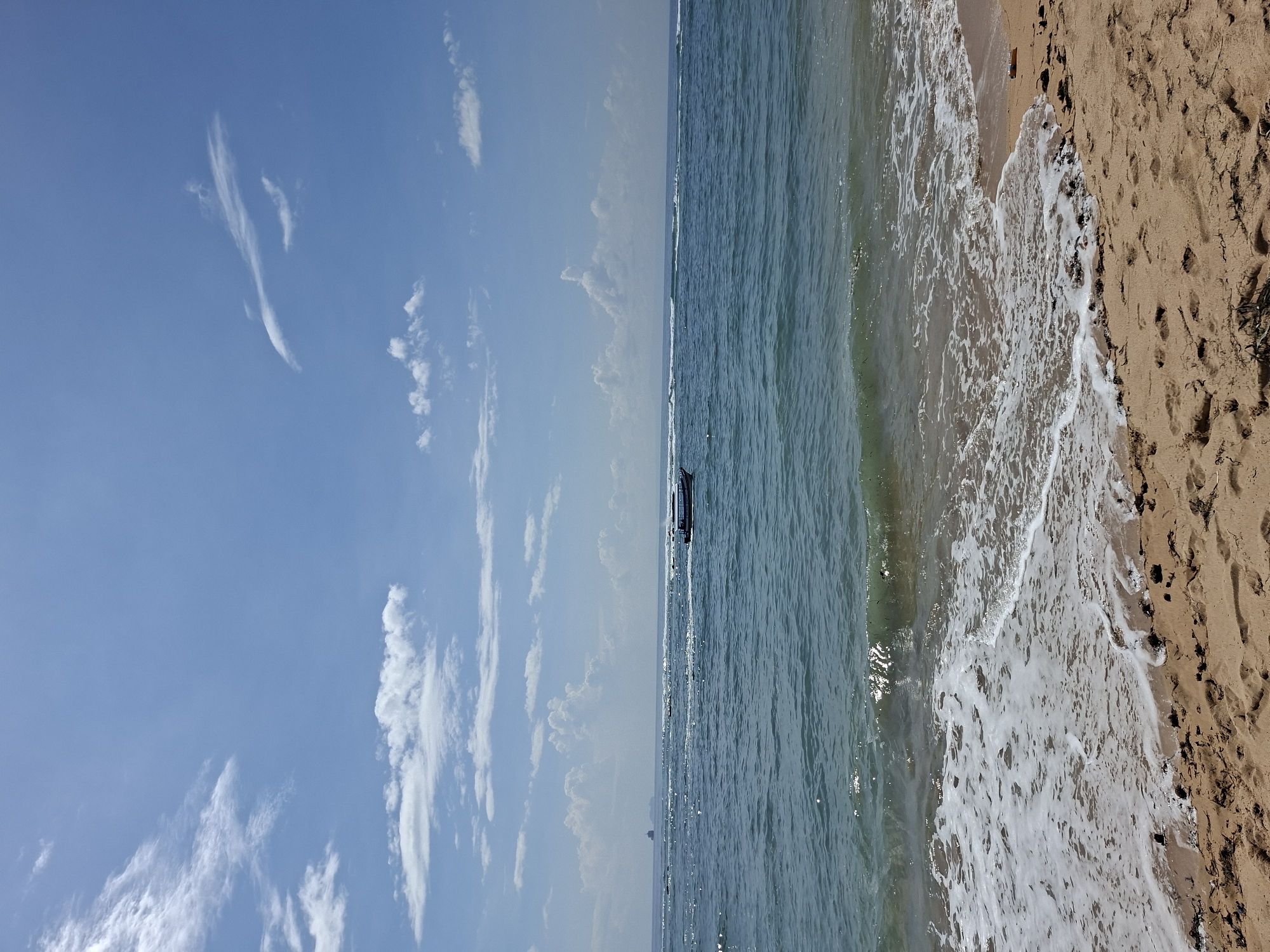 This vibrant image depicts a serene coastal scene in Kuta Selatan, Indonesia, under a clear, bright sky. The composition is horizontally divided into three main sections: the sky, the ocean, and the sandy beach.
The upper portion of the frame is dominated by a brilliant blue sky, adorned with scattered, wispy white clouds that stretch across the expanse. Below the sky, the expansive ocean shimmers with varying shades of blue and green, reflecting the bright daylight. A small, dark-colored boat is visible in the middle distance, positioned on the calm waters near the horizon line.
Closer to the foreground, gentle white foamy waves can be seen breaking and receding onto the shore, creating a dynamic edge between the water and land. The lower section of the image features the warm, golden-brown sand of the beach. Numerous footprints are imprinted into the sand, indicating recent activity, and small patches of darker debris, possibly seaweed or vegetation, are visible near the waterline.
The scene suggests a peaceful daytime setting, likely morning or afternoon, given the clear skies and strong natural light. There is no visible text in the image. The overall impression is one of natural beauty and tranquility at a tropical beach.