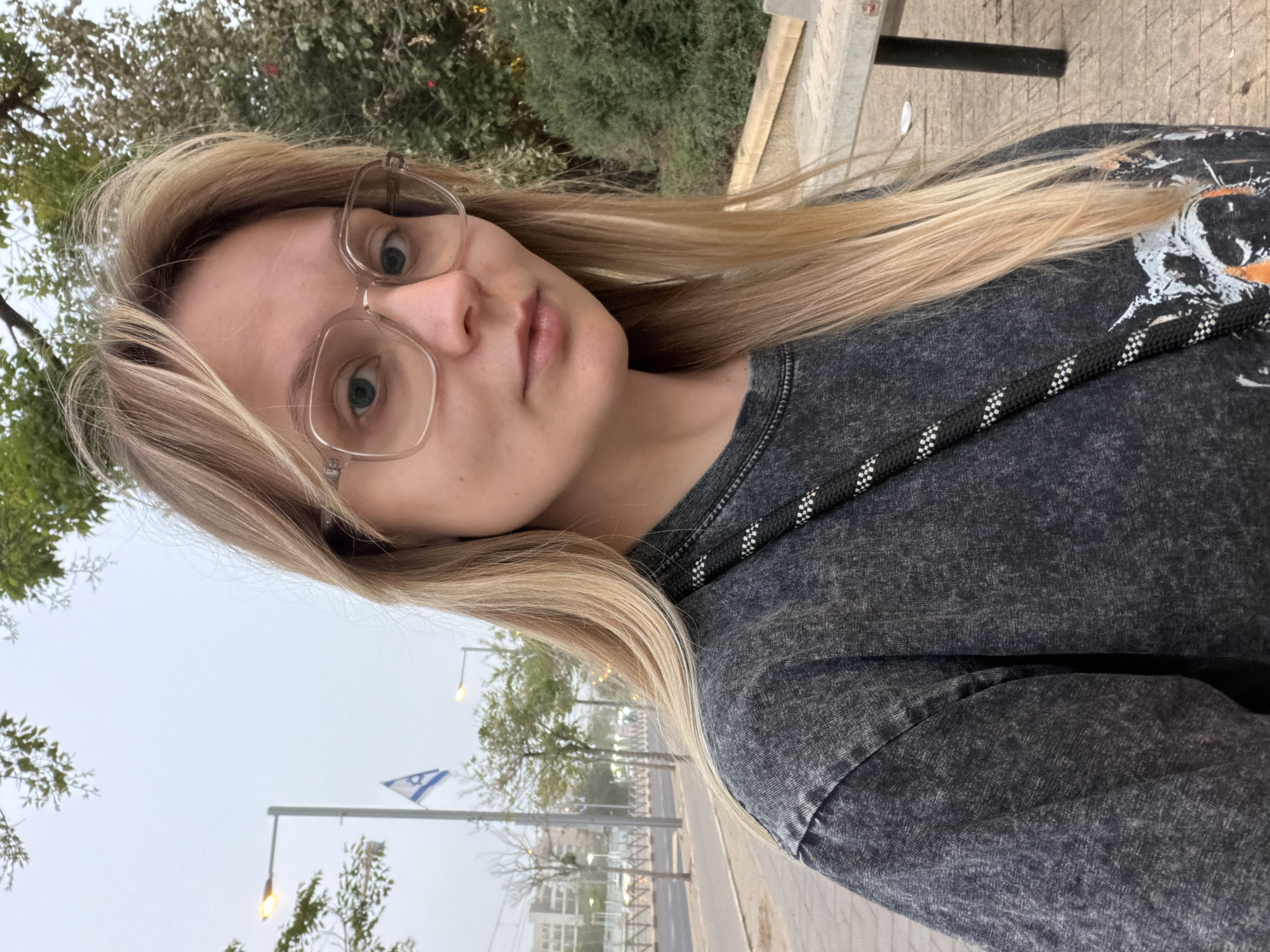 A medium-shot selfie depicts a fair-skinned individual with blonde hair and glasses. They wear a dark gray garment. The background suggests an urban environment, with streetlights and a building visible. A blue and white flag, the Flag of Israel, is recognizable. The presence of the flag and architecture indicate the location is most likely in the city of Tel-Aviv, Israel.