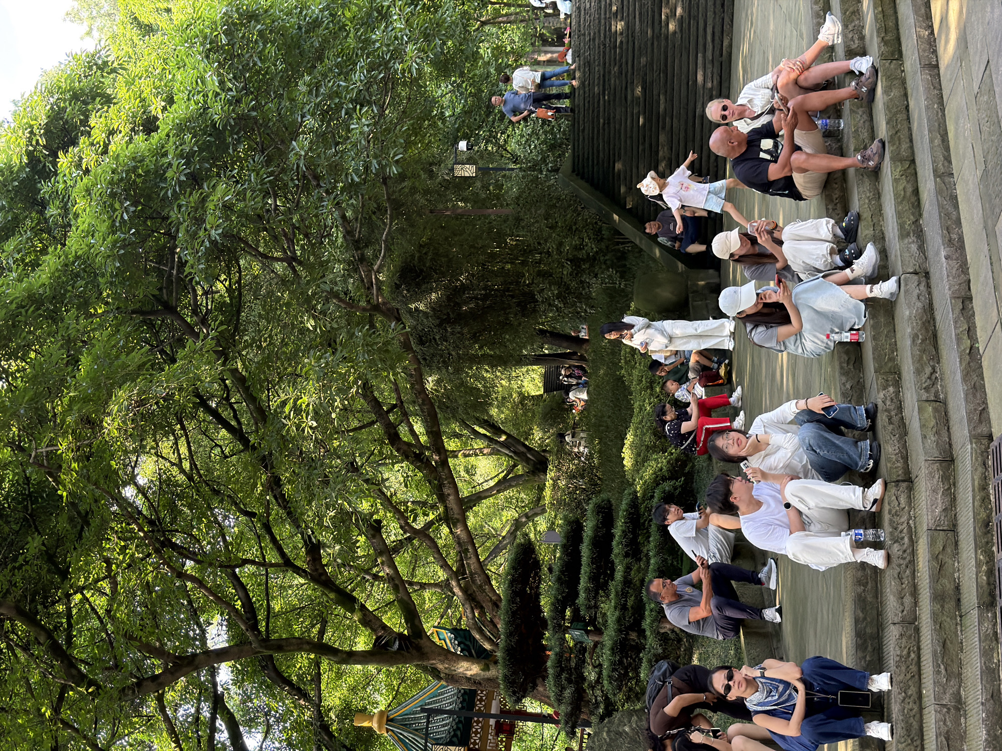 The image depicts a group of people in a park setting, with lush green trees and stone steps dominating the scene. In the foreground, several individuals are seated on stone steps, some looking at their phones, others conversing. A woman in blue overalls and a patterned scarf is seated on the left, looking to her left. Next to her, two individuals, one in a white t-shirt and the other in a white top and jeans, are seated, with the latter looking at her phone. Further to the right, a man in a black t-shirt and shorts is seated with a woman in a white striped shirt and sunglasses. In the background, a set of wide stone stairs ascends to the right, with several people walking up and standing on them. A small child in a hat is visible on the stairs. A traditional Chinese-style pavilion with a green and yellow roof is visible to the left, partially obscured by trees. The environment suggests a public park with landscaped gardens and architectural elements. The location is specified as Yuzhong District, China.