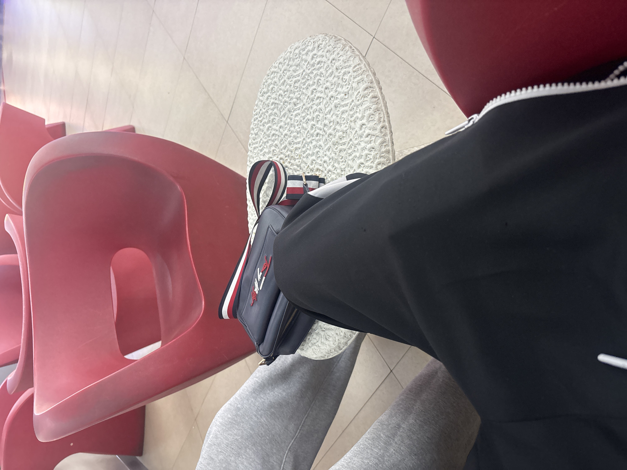 This image presents an overhead, angled view of a person in what appears to be an indoor public waiting area. The setting features several bright red, molded plastic chairs, common in places like airports, bus stations, or clinics. A tiled, light-colored floor, possibly off-white or light beige, extends across the visible space.
The primary subject is a person, with their lower body in grey sweatpants and a black long-sleeved top visible. A notable object is a small, dark blue messenger bag or crossbody bag, prominently placed in the center of the frame. This bag rests on a white, highly textured surface, which appears to be the sole of a shoe worn by the person. The bag features a distinctive red, white, and blue striped strap and a clear "TOMMY HILFIGER" logo in white script with the brand's iconic flag emblem.
The person appears to be seated or resting in one of the waiting area chairs. There are no overt activities taking place beyond this state of rest or waiting. The lighting suggests an indoor environment, making the time of day and weather indiscernible. No specific emotional expressions are visible from the partial view of the person. Given the provided context, the scene is situated in Yerevan, Armenia, although the visual cues within the image are generic to public waiting areas.