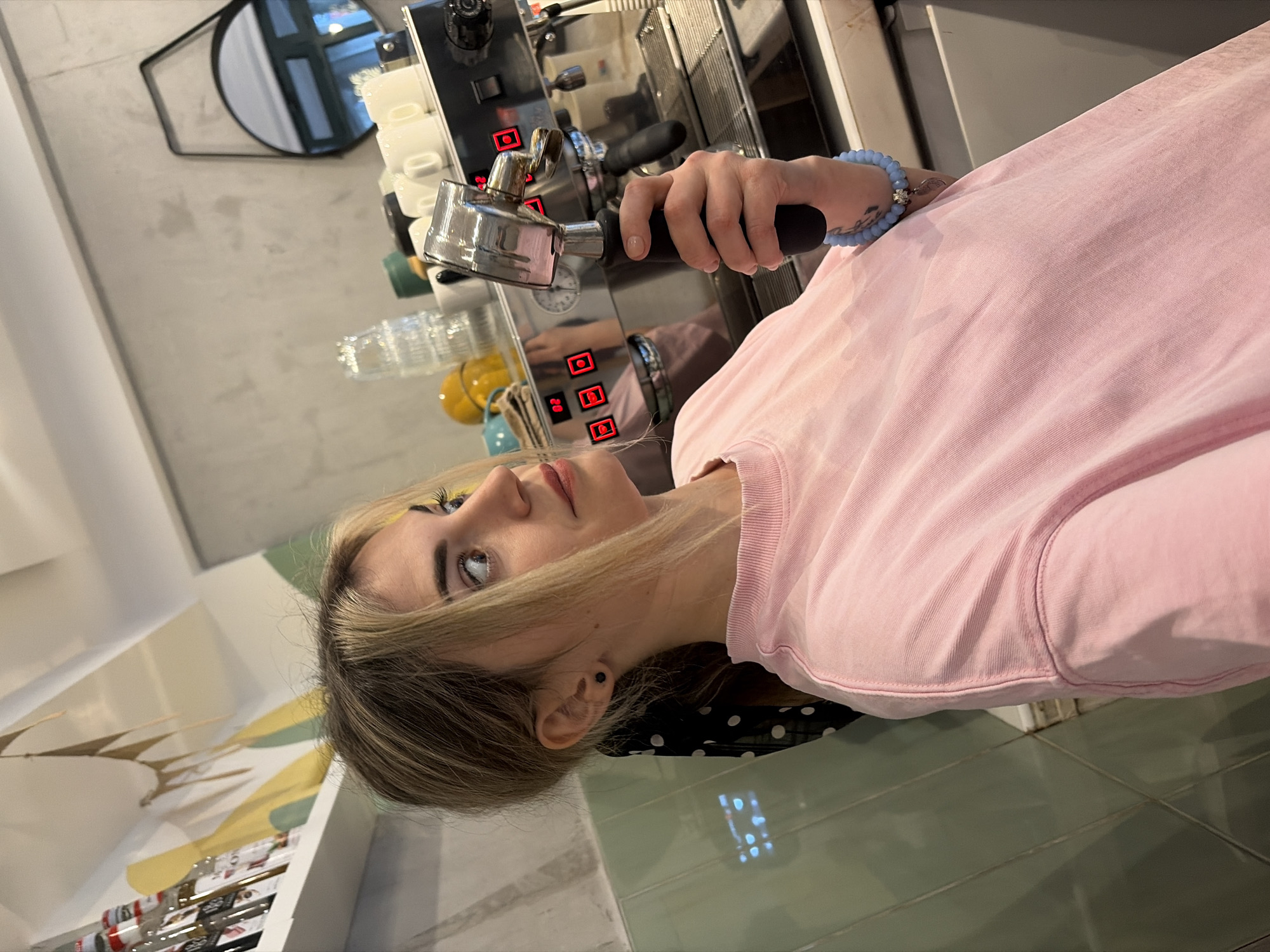 The image displays a person, appearing to be a young woman with blonde hair, actively engaged with a commercial-style espresso machine in what looks like a cafe or kitchen environment. The image is oriented sideways, with the person's head towards the lower left and their body extending towards the upper right of the frame.
The woman has light blonde hair, slightly darker at the roots, and is wearing a plain, light pink long-sleeved shirt. Her left eye is clearly visible, featuring prominent yellow eyeshadow and dark eyeliner, as she gazes upwards and slightly to her right. A small, black stud earring is visible in her left ear, and her lips have a soft, natural pink tint.
Her right hand is holding a metallic portafilter by its black handle. The portafilter is connected to, or is about to be connected to, a robust silver-toned espresso machine. The machine has a series of red illuminated square displays or buttons, two of which clearly show the numbers "20" and "22". White ceramic coffee cups are stacked neatly on top of the espresso machine. Around the woman's right wrist, a light blue beaded bracelet with a small metallic charm is visible, and a partial tattoo can be seen on her forearm beneath the bracelet.
The background features a light gray wall. Hanging on this wall is a circular mirror with a black frame, reflecting what appears to be a window or doorway, showing vertical lines and bright light from outside or another room. Below the mirror, several clear glasses or bottles are present, along with a yellow, possibly ceramic, jug or container.
Beneath the espresso machine and extending towards the foreground, there is a counter or floor area made of greenish-gray tiles. A section of black fabric with white polka dots is visible near the woman's neck and shoulder. To the far left of the frame, shelves stocked with various packaged items or bottles are partially visible. The scene is brightly lit, suggesting either daytime or well-lit indoor conditions.
