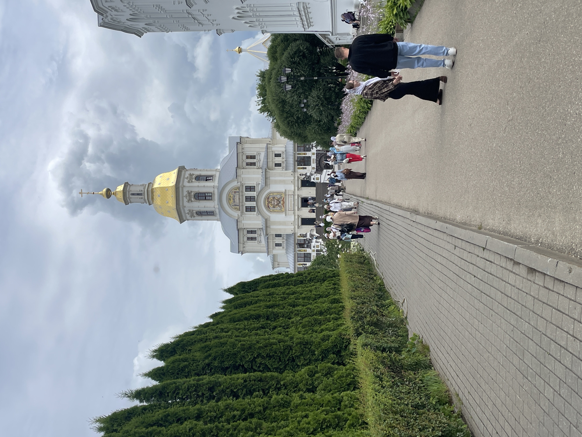 The image depicts a view of the Holy Trinity Cathedral in Diveyevo, Russia, from a paved walkway. The cathedral, with its prominent golden dome and cross, is situated at the end of the path. A dense line of manicured evergreen trees runs along the left side of the walkway, separating it from a lower hedge. Several people are walking along the path towards the cathedral. Some are dressed in traditional modest clothing, with women wearing headscarves. In the foreground, a couple walks away from the camera. The sky is overcast with dramatic clouds. The right side of the image shows a white building with arched windows, likely part of the monastery complex. Flowers are visible in a garden bed along the right edge of the walkway.