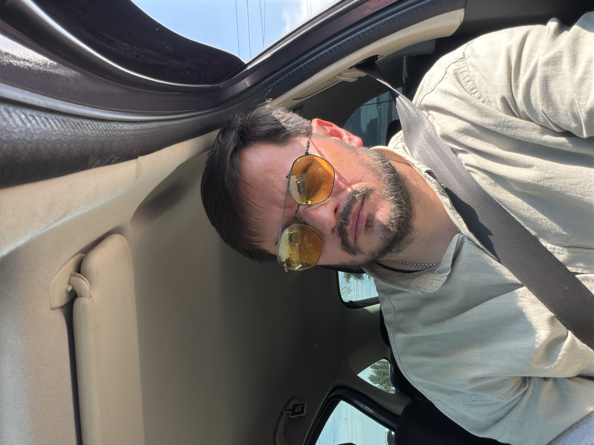 A man is seen from the chest up, sitting in the driver's seat of a car. He is wearing a light-colored shirt, a silver chain, and sunglasses with orange lenses. The man has short dark hair and a beard. The interior of the car is visible, including the sun visor and the seatbelt. The window on the right side of the car is open, showing a bright sky and some power lines in the distance. The lighting suggests it is daytime. There are no discernible landmarks or details that would indicate the specific location within the city of Donetsk, Ukraine.