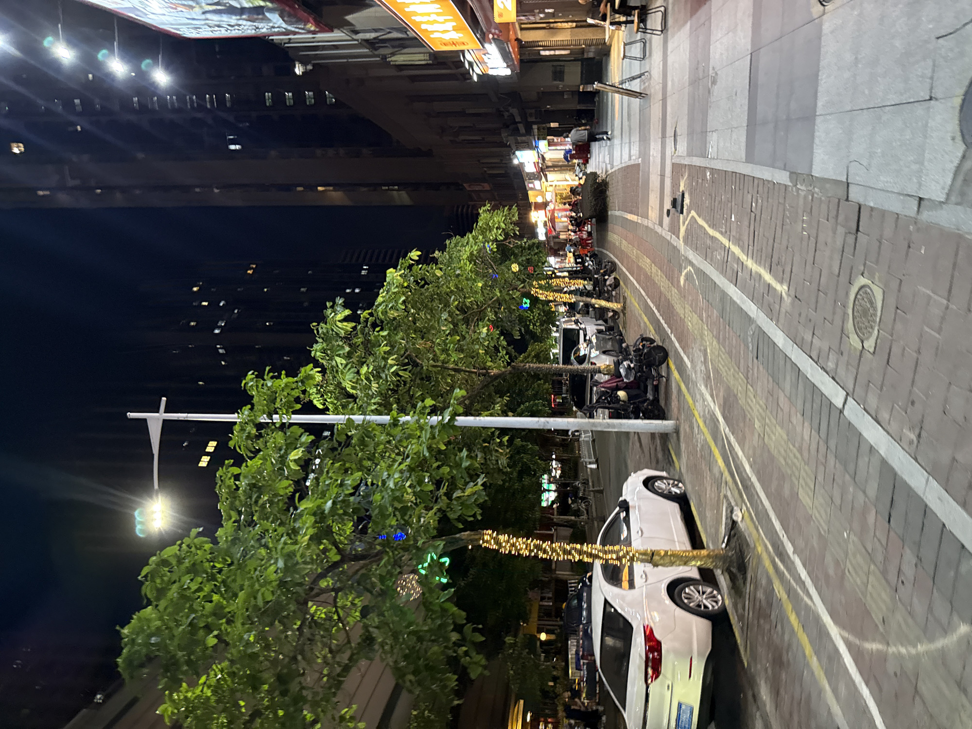 The image depicts a nighttime street scene in Haizhu District, China. Tall buildings line the right side of a narrow street, illuminated by various lights from windows and signs. A sidewalk runs along the buildings, with several tables and chairs set up outside what appear to be restaurants or shops. People are visible walking and sitting on the sidewalk. The street itself is paved with bricks and features a bike lane marked by yellow and white lines. Several trees are planted along the street, decorated with string lights. A white sedan is parked on the left side of the street, and various motorcycles and scooters are parked along the curb and in the bike lane. A traffic light is visible in the upper left corner of the image, above the trees. The overall impression is of a busy, urban environment at night.