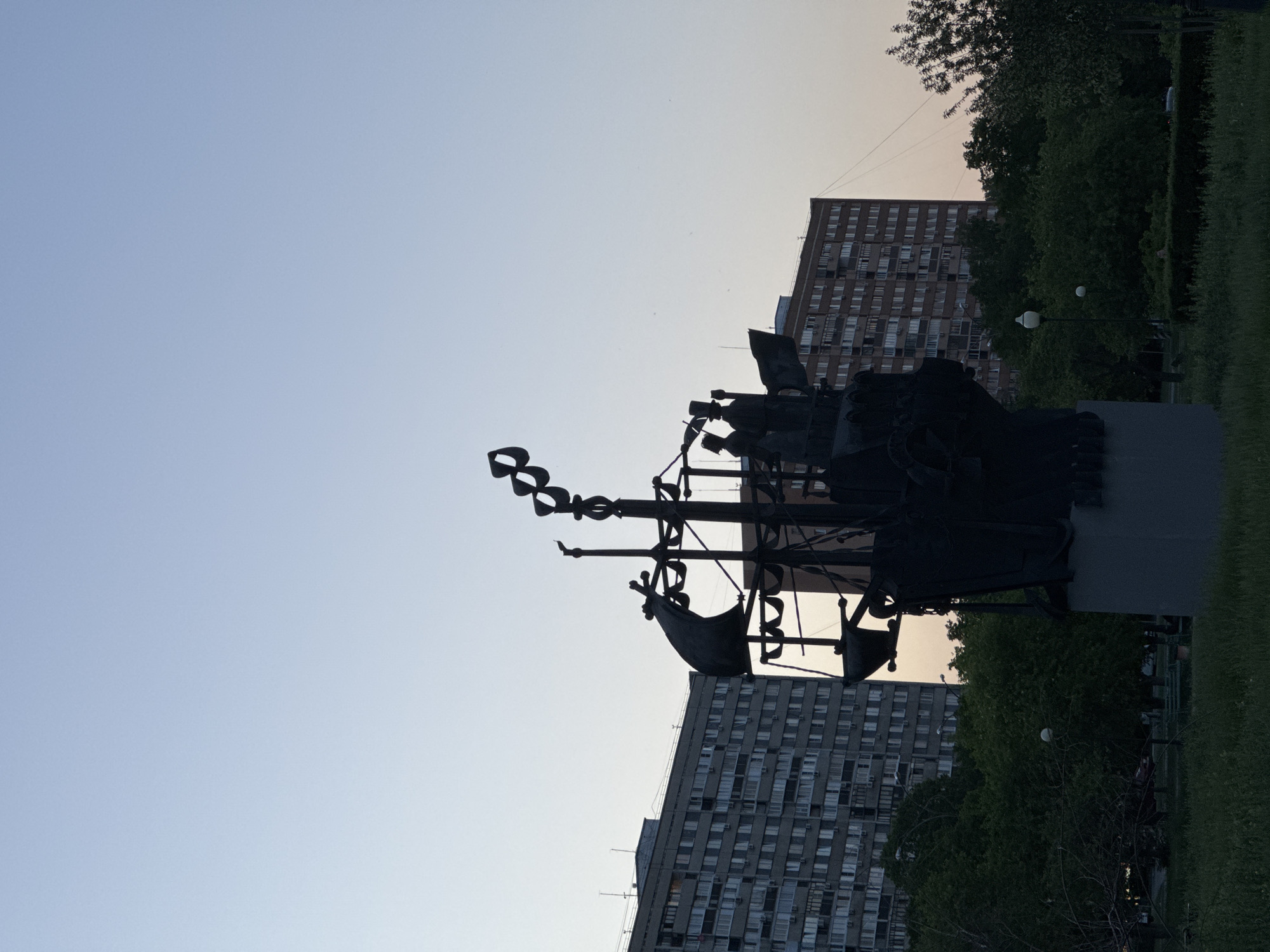 Here is a description of the image:
The image features a dark silhouette of a modern sculpture in a park setting in Unknown, Unknown. The sculpture includes abstract shapes and figures, including what appears to be stylized boats or ships. Behind the sculpture are two large residential buildings with numerous windows. A line of trees and green grass runs along the right side of the image. The sky above the sculpture is light blue, transitioning to a pale orange near the horizon. The photograph is oriented vertically.