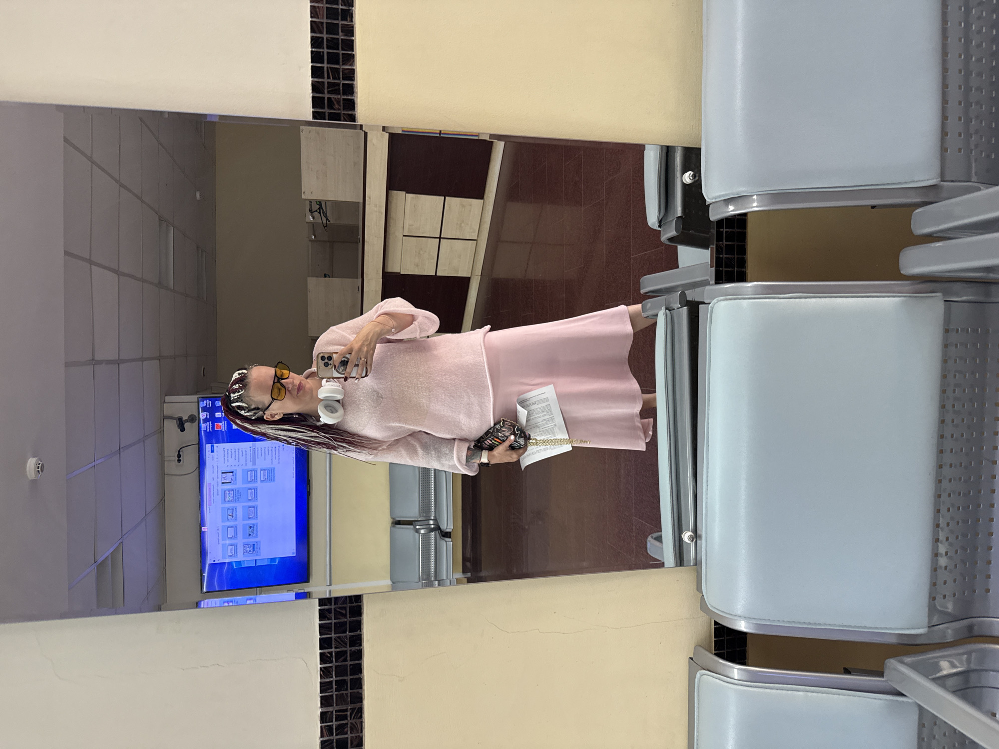 A mirror selfie captures a woman in an indoor public setting, likely a waiting area in Moscow, Russia. The image is presented rotated 90 degrees clockwise.
The subject, a woman, is reflected in a large mirror. She wears a light pink long-sleeved top and a matching light pink skirt. Her hair is styled in long, multi-colored braids or dreadlocks. She sports yellow-tinted sunglasses and white over-ear headphones. In her left hand, she holds a white smartphone, positioned as if taking the selfie. Her right hand holds a document or papers and a small, patterned clutch bag. Rings are visible on her left hand.
The background, seen in the reflection, features a large flat-screen television mounted on a wall, displaying what appears to be a Windows file browser or image gallery interface. Above the TV, a white circular smoke detector is visible on the light-colored, tiled ceiling. The room has light beige/yellow walls, with sections of dark brown square mosaic tiles. The floor is covered in dark red/brown tiles. On the right side of the reflection, rows of light blue upholstered waiting chairs with metal frames are visible, indicating a public waiting area or lounge.
The scene depicts a calm, well-lit interior. No specific legible text is discernible on the television screen or the documents held by the woman.