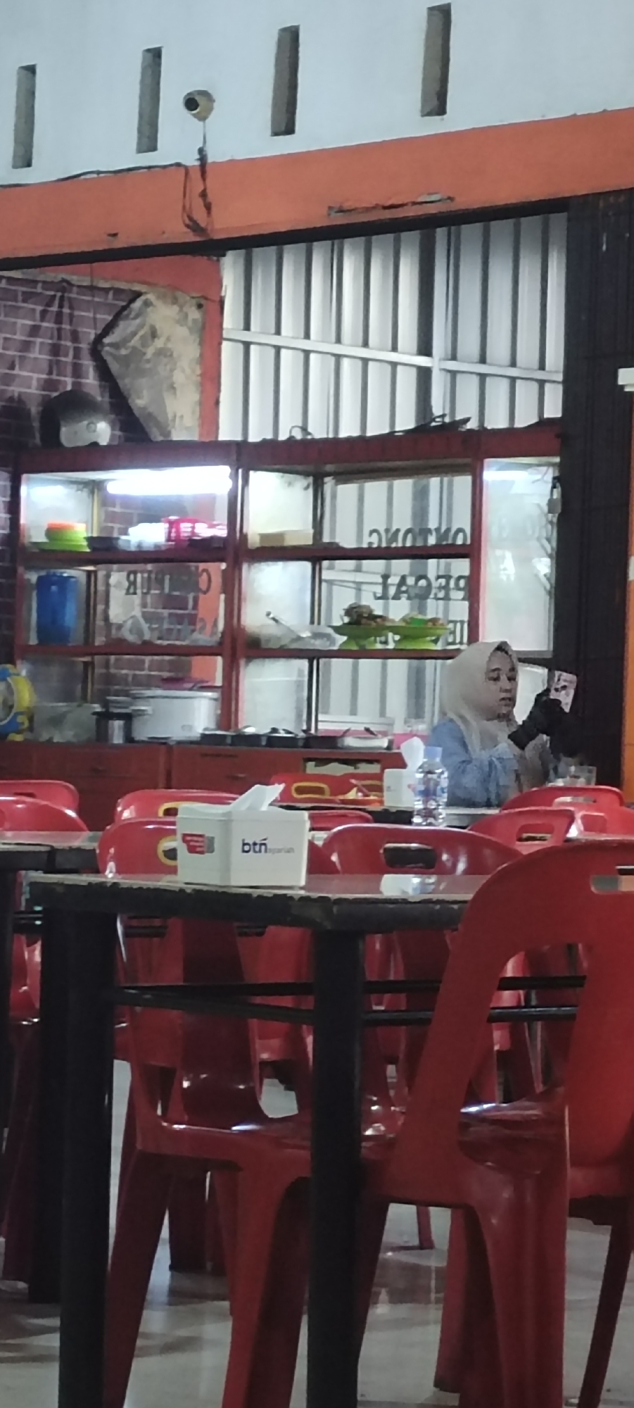 The image shows the interior of a food stall or small restaurant in Lambaro, Indonesia. In the background, a woman wearing a light blue headscarf and a light blue shirt is seated at a table, holding a small object, possibly a phone or card. She is looking towards the right side of the frame. Behind her, a glass display case contains various food items and containers. Red chairs are arranged around tables in the foreground, with a tissue box on one of the tables bearing the "btn syariah" logo. The walls have a textured pattern and an orange trim runs along the top of the display area. The flooring appears to be concrete.