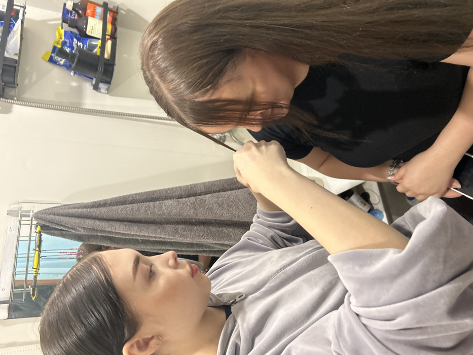 In an indoor setting, likely a bathroom or salon in Chaikovsky, Russia, two women are engaged in a hair-styling activity. The woman on the left, with dark hair pulled back and wearing a light gray zippered hoodie, is observed in profile, focused on the task. Her right hand holds a thin strand of hair from the top-front section of the second woman's head. The second woman, positioned on the right and facing away from the camera, has long, medium brown hair and wears a black t-shirt. Her head is tilted slightly forward and to the left. Her right hand is visible, holding a white object below the frame. The background features a rectangular mirror with a light frame, a gray towel hanging from a rack, and a shower hose. To the right, two black wall-mounted shelves hold assorted bottles, pouches, and containers against a white wall.