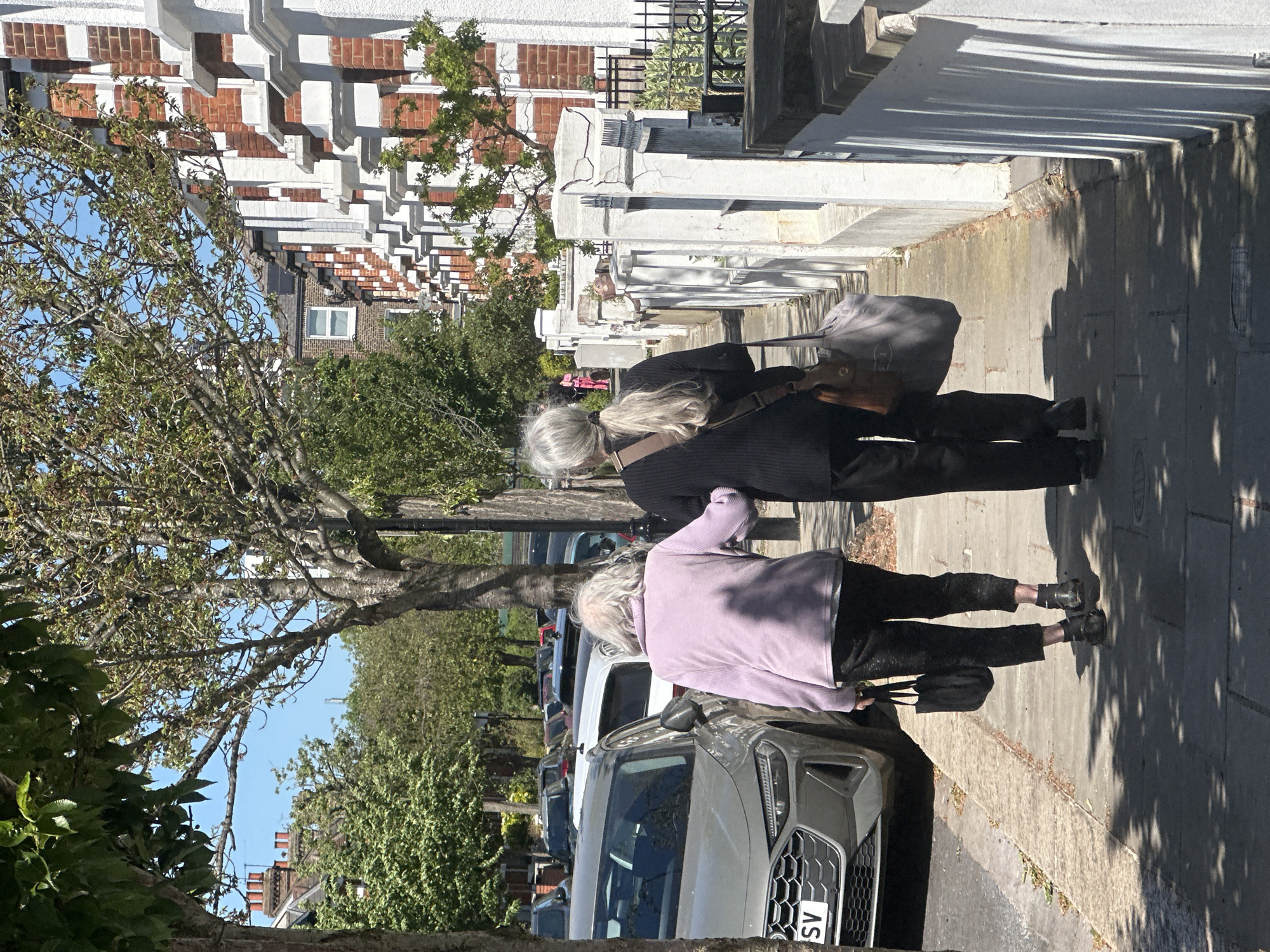 This image, rotated 90 degrees counter-clockwise from its intended orientation, depicts a bright, sunny day on a residential street in what appears to be London, United Kingdom. Two individuals, both women with grey hair, are walking away from the camera on a paved sidewalk.
In the foreground, one woman wears a light purple long-sleeved top and dark trousers, carrying a dark handbag on her arm. Her short, light grey hair is visible. Behind her, and slightly closer to the buildings, walks another woman with longer, light grey hair tied back in a ponytail. She is dressed in a dark top and dark trousers, and carries a grey shoulder bag with a brown strap.
A line of parked cars extends along the street to their left, with a light grey SUV prominently featured in the mid-ground. The visible license plate on the SUV displays the letters "SV". The street is lined with numerous mature trees, some with dense green foliage and others with more visible branches, casting distinct long shadows across the sidewalk.
To the women's right, a row of multi-story terraced houses stretches into the distance. These buildings feature classic red brick facades, complemented by white architectural details such as bay windows, cornices, and decorative trim, typical of London's urban landscape. The sky is clear and blue, and the strong sunlight indicates either a morning or late afternoon setting.