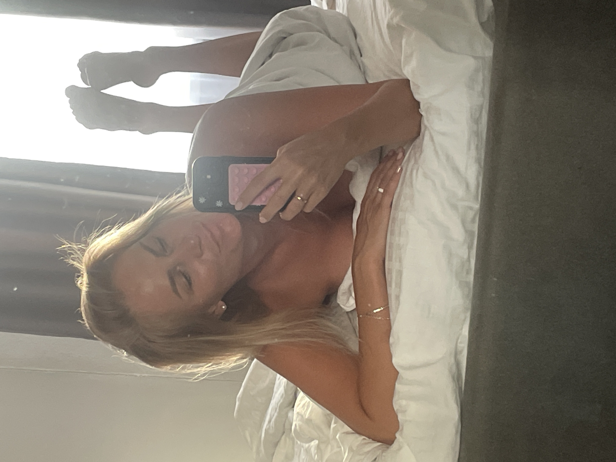 A woman is lying on a bed in a room, taking a selfie. She is wrapped in a white sheet, with her legs in the air. She has blonde hair and is wearing a ring on her finger. The room appears to be a bedroom, with a white duvet and pillows. There is a window in the background, with curtains. The lighting suggests it is daytime. There is no information in the image to indicate the location in the city of Baranovka, Russia.