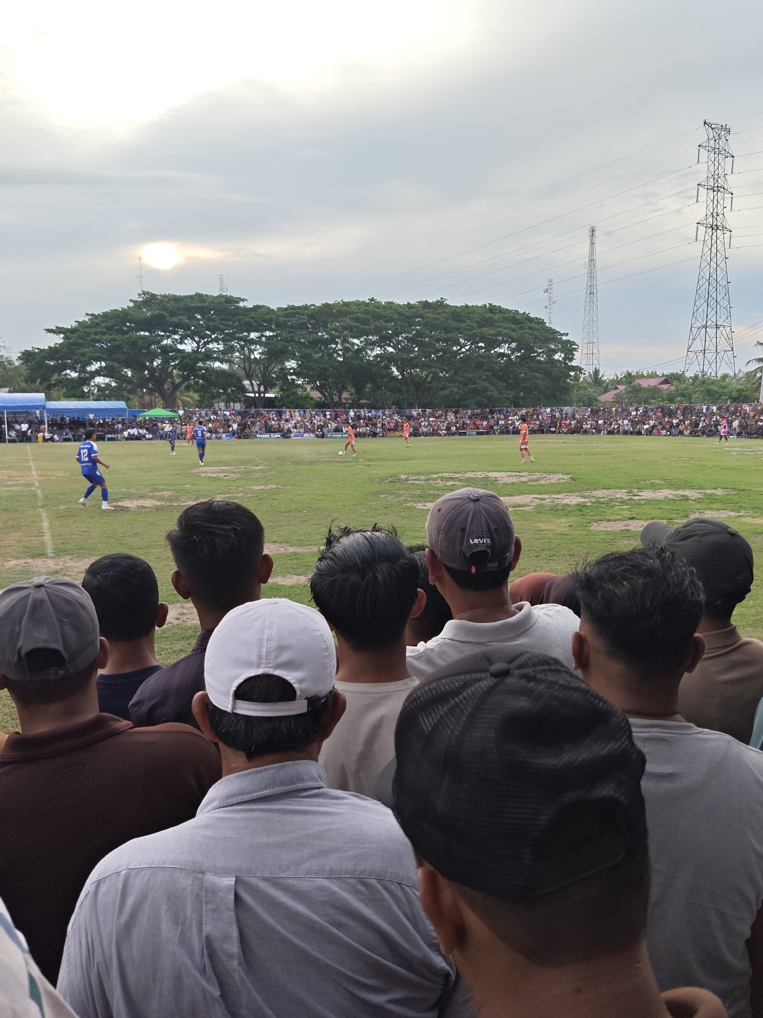The image depicts a crowded outdoor football match viewed from behind a section of spectators. The scene is set on a grassy field with white markings. Two teams are visible on the field: one in blue jerseys with the number 12 prominent, and another in orange jerseys. Numerous spectators are gathered in bleachers and around the field, some under tents. In the background, large trees and several tall electrical transmission towers are visible against a cloudy sky with the sun partially obscured. The foreground shows the backs of several spectators wearing hats and casual clothing. The overall impression is of a lively community event taking place in a semi-rural setting.