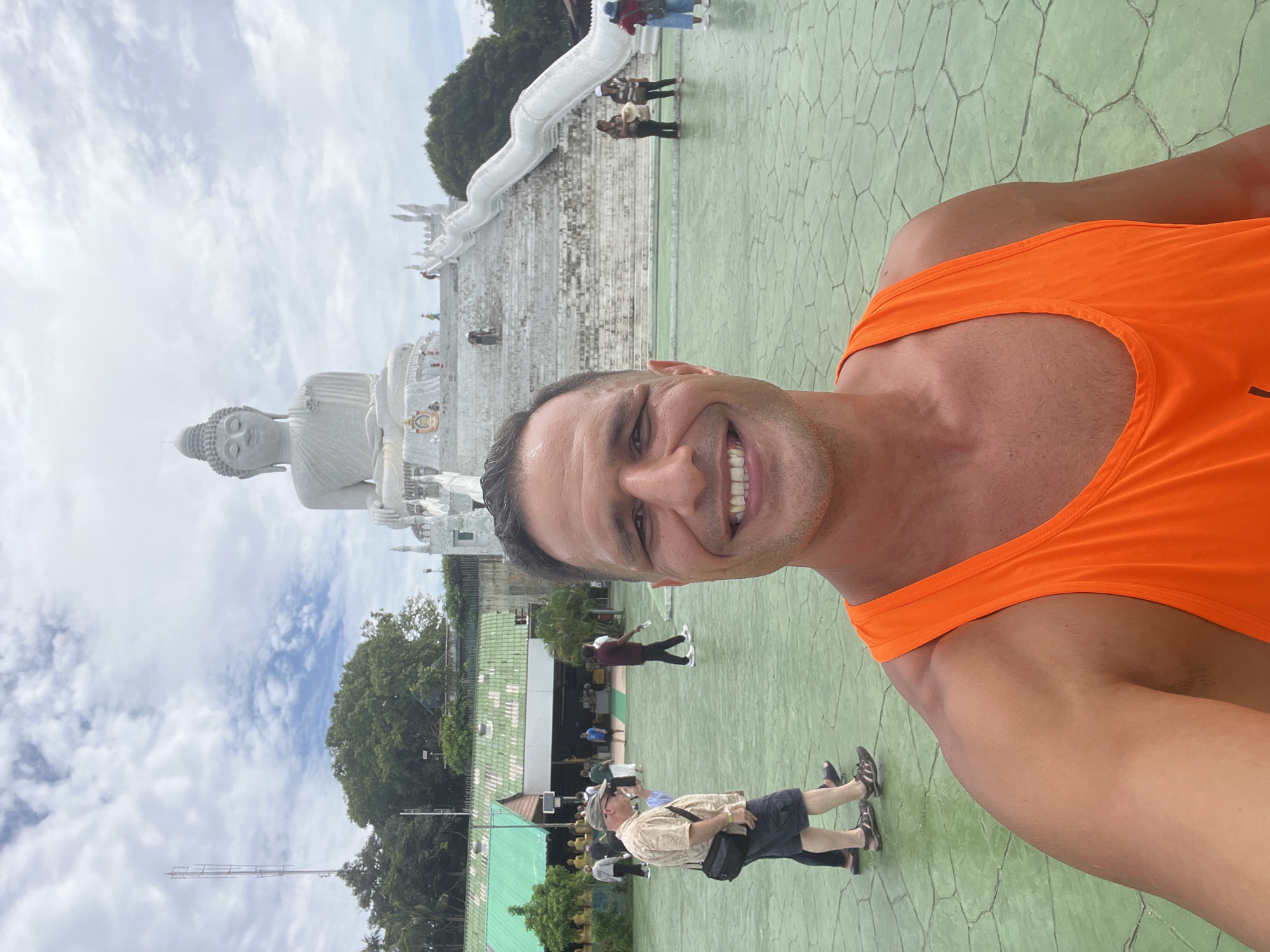 A man in an orange tank top smiles at the camera in a selfie. Behind him, a large white Buddha statue sits atop a white tiered structure with stairs leading up to it. Several other people are visible in the background, walking around the plaza. The ground is a light green, textured pavement. The location is the Big Buddha of Phuket in Karon, Thailand, a prominent landmark. The sky is partly cloudy.