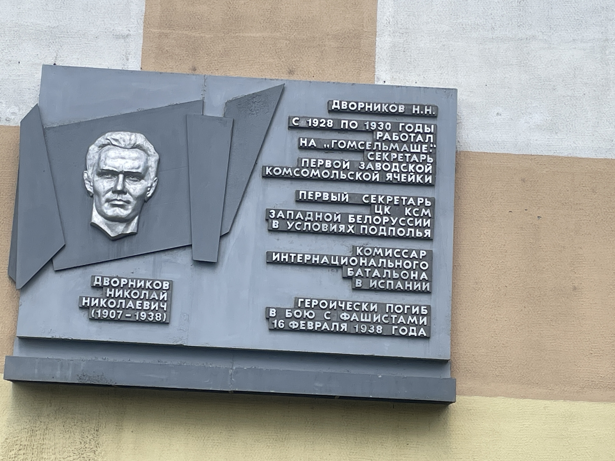 The image shows a memorial plaque on a building in Homyel, Belarus, dedicated to Dvoornikov Nikolai Nikolaevich (1907-1938). The plaque features a bas-relief portrait of a man and text in Russian. The text details his career, including his work at "Gomselmash" from 1928 to 1930, his role as first secretary of the Gomel regional Komsomol committee, and his service as commissar of the international battalion in Spain, where he died in battle against fascists on February 16, 1938. The building's exterior wall is visible, painted in a beige and off-white color.
