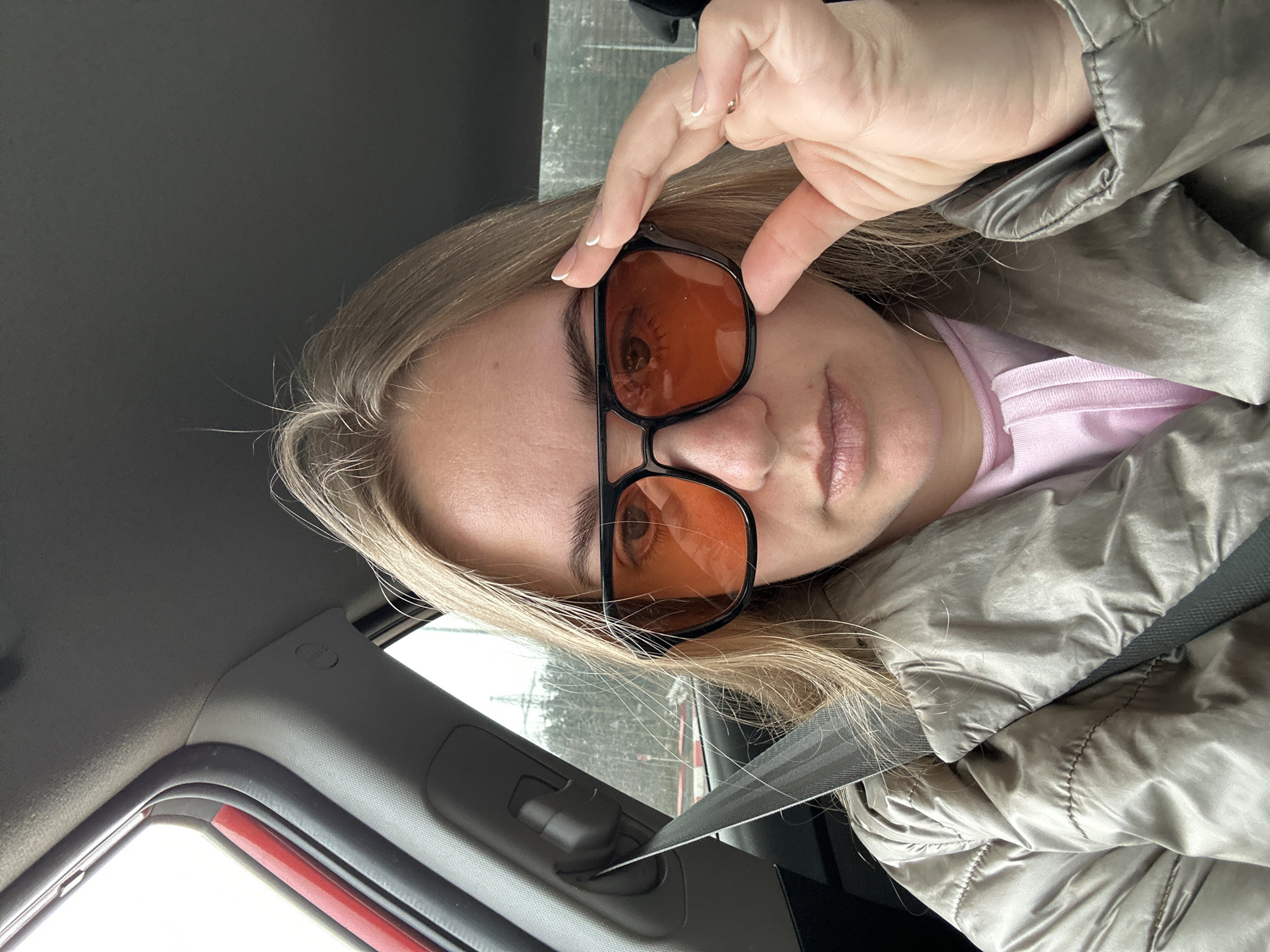 A woman is in a car, wearing orange-tinted sunglasses, a pink top, and a shiny, grey jacket with the seatbelt fastened. She is holding the top of her sunglasses with her right hand. The interior of the car is visible, with a red window frame on the left side of the image. The location in the city of Chaikovsky, Russia is not discernible from the image.