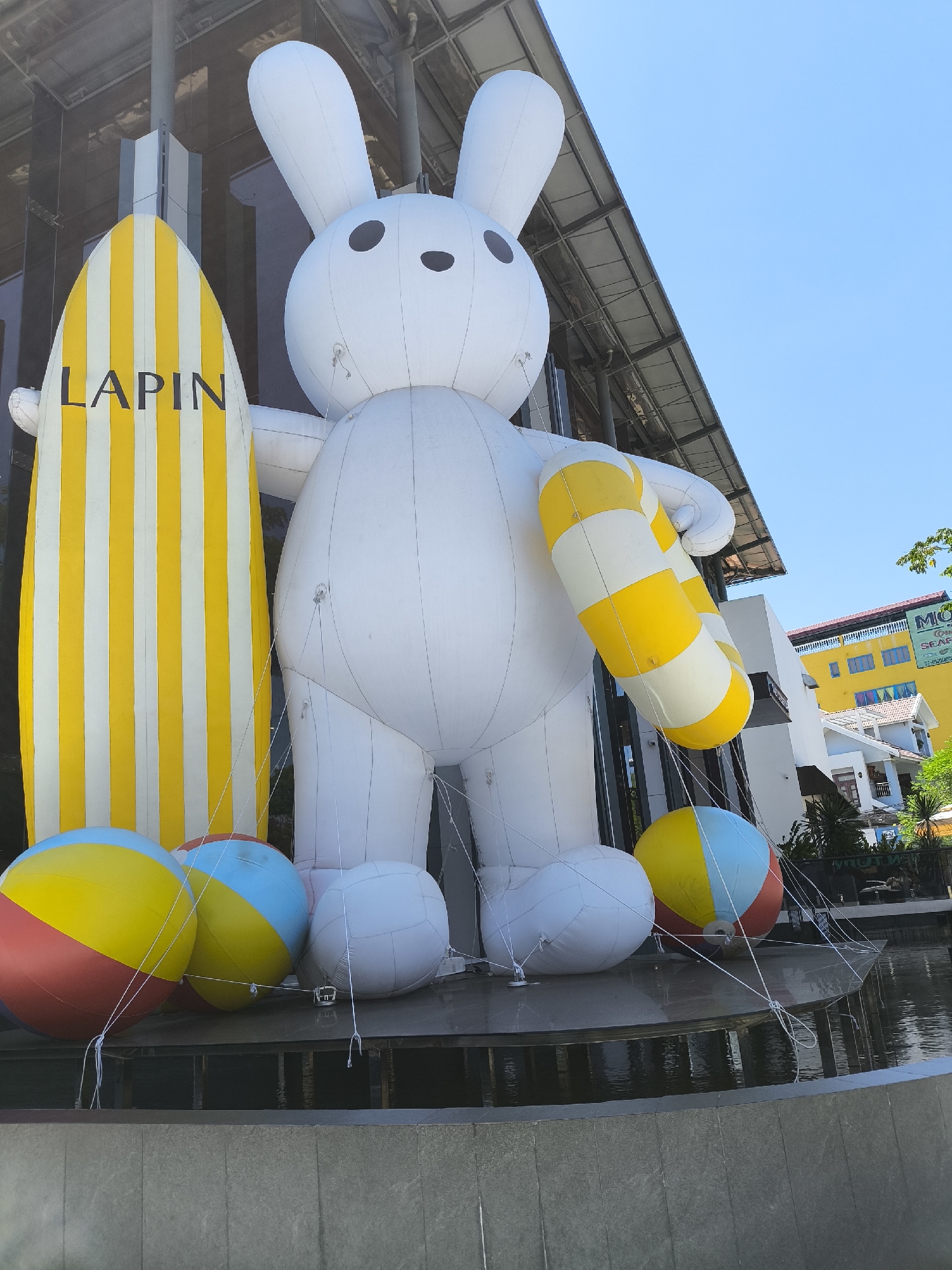 The image depicts a large inflatable white bunny character standing outside a building in Đà Nẵng, Vietnam. The bunny is holding a yellow and white striped inflatable ring. Next to the bunny is a surfboard with yellow and white vertical stripes, bearing the text "LAPIN". In front of the bunny and surfboard are several large inflatable balls, one with red, yellow, and blue segments, and others that are white. The scene appears to be set in an outdoor plaza or walkway with a body of water in the foreground. The building behind the display has a modern architectural style with large glass windows and a metal roof structure. In the background, other buildings are visible, including a yellow building with signage that is partially obscured but appears to include "MCI". The sky is clear and blue.