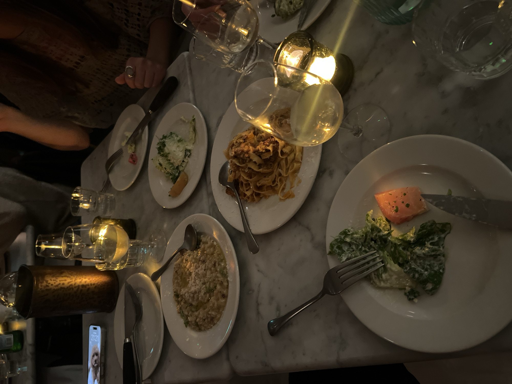 The image depicts a table setting with multiple plates of food, including pasta bolognese, risotto, and a salad with a slice of salmon. There are also glasses of white wine and a candle. A person's hand is visible near a plate of salad, and a cell phone with an image of a dog is lying on the table. The setting appears to be indoors, likely a restaurant. The location is possibly in the city of London, United Kingdom.