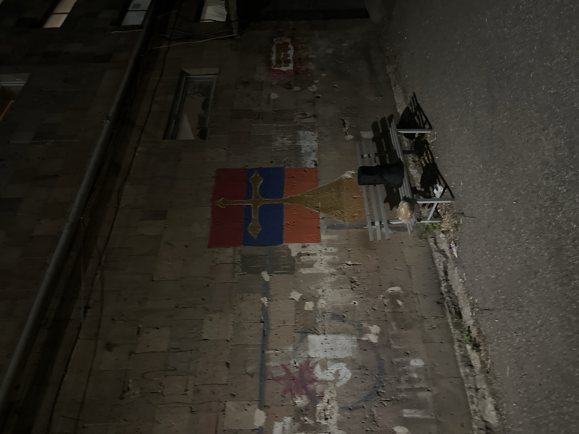 The image depicts an alleyway in Yerevan, Armenia. On the left is a building with exposed pipes, a window, and peeling brickwork. An Armenian flag is painted onto the wall. There are other tags or patches of graffiti on the wall, and debris litters the alleyway. A metal bench with a dark bag on it is situated in the foreground of the alleyway. The alleyway is poorly lit.