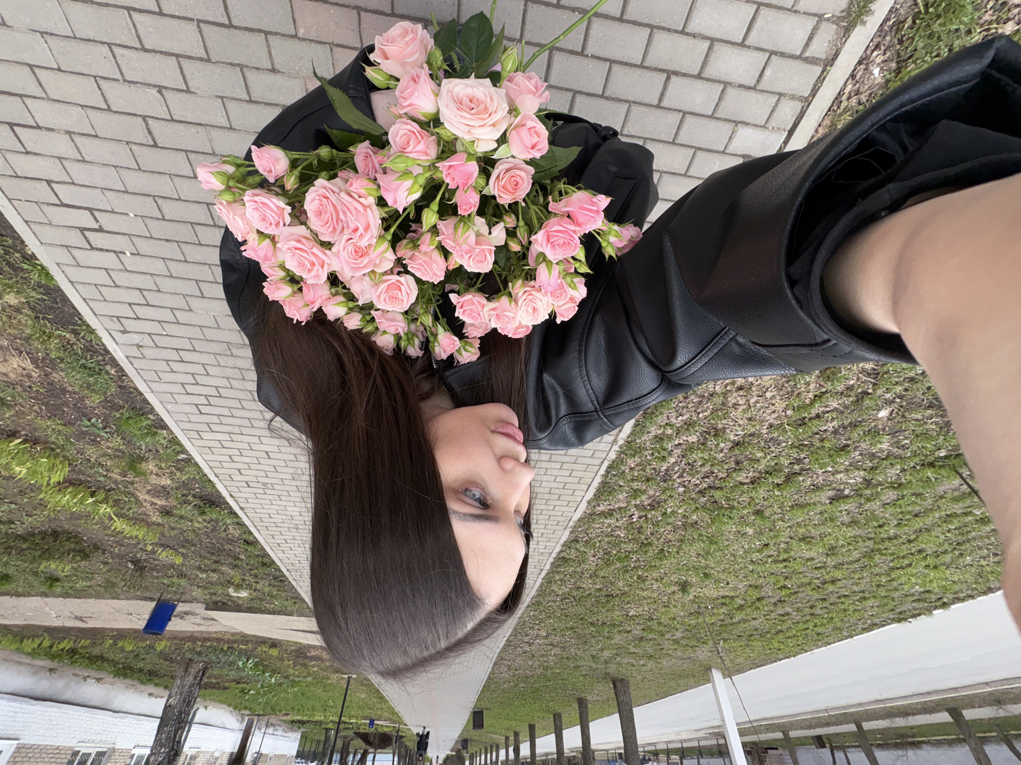 A young woman is depicted from an overhead perspective, holding a large bouquet of pink spray roses. Her long, dark brown hair cascades down her side, partially covering her shoulder. Her face is shown in profile, with light-colored eyes looking gently towards the right (her right). She is wearing a black leather jacket.
The bouquet of roses is a prominent feature, consisting of numerous light pink blooms and buds, tightly clustered together. The flowers appear fresh and vibrant.
The setting is outdoors, likely a paved pedestrian area or park in Chaikovsky, Russia. The ground beneath her is a light gray brick-patterned pathway. To the side of the path, there is an expanse of green grass mixed with some drier patches of earth. In the background, faint details of distant structures, possibly buildings or fences, and sparse trees or posts can be discerned along what appears to be another path or road.
The lighting indicates daytime conditions, possibly overcast, given the soft and even illumination without strong shadows. The woman's expression is calm and neutral. There is no visible text in the image.