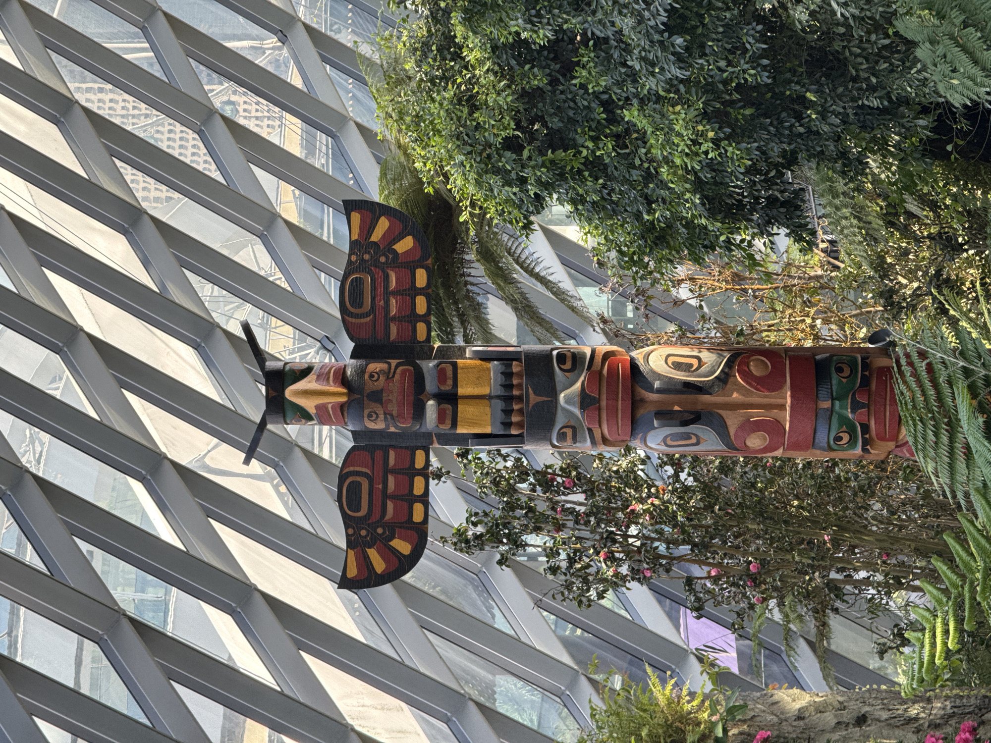 The image features a totem pole set against a backdrop of a glass and metal lattice structure, possibly a greenhouse or conservatory. The totem pole is prominently displayed, with detailed carvings in red, black, yellow, and other colors. The background includes lush greenery and trees, adding a natural element to the scene. The location appears to be an indoor botanical garden or similar structure.