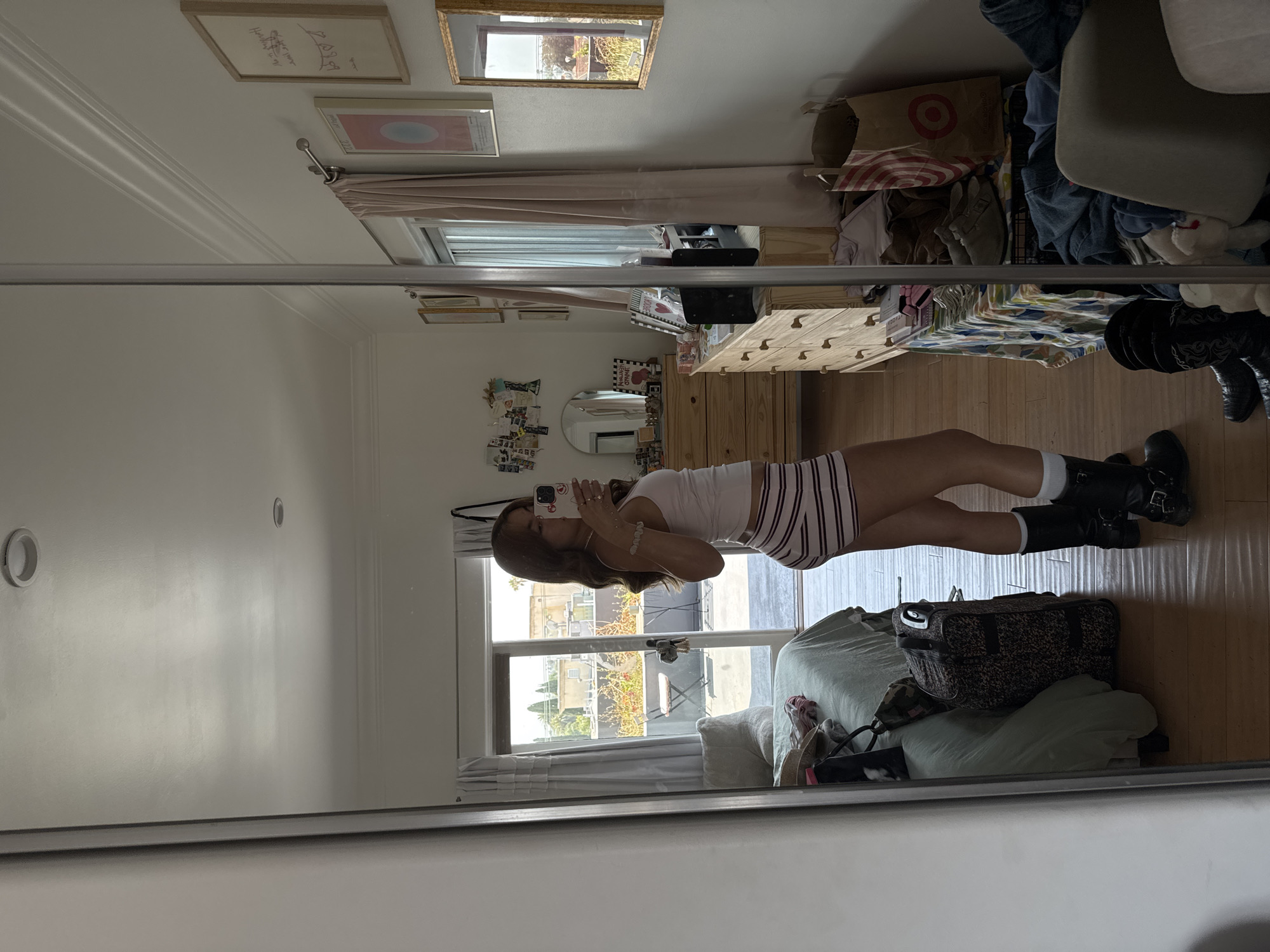 A reflection in a closet mirror shows a person wearing a striped outfit, white socks, and black boots standing in what appears to be a bedroom. The person is holding a phone and taking a picture of their reflection. Behind the person, there is a bed with a green comforter and pillows, and a suitcase on top of the bed. A window with white curtains is visible, and outside the window, there is a view of buildings and greenery, indicative of an urban environment. Based on the architectural styles and vegetation, it is possible that the location is in the city of Los Angeles, United States of America. The room also contains a wooden dresser, various decorations, and a Target shopping bag on a shelf.
