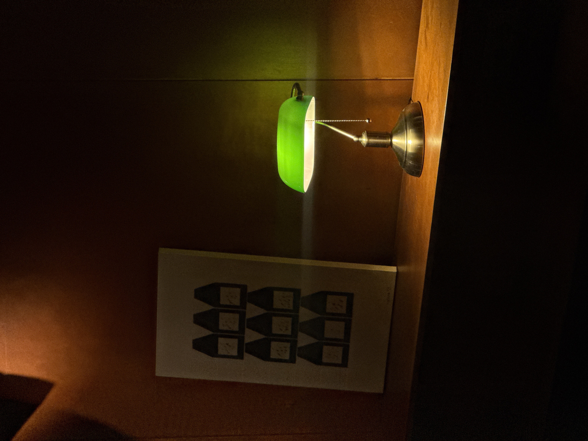 A green banker's lamp with a brass base illuminates a wooden shelf in a dimly lit room. To the left of the lamp, a framed print depicting nine identical, dark-silhouetted bottles with white labels is leaning against a textured, reddish-brown wall. The lamp casts a bright, green glow on the wall behind it, creating a gradient of light. The room appears to be a residential or office space, possibly a home study or library. There are no people or animals present in the scene. The overall atmosphere is quiet and contemplative, with the light from the lamp drawing attention to the artwork and the immediate surroundings. The image quality is consistent with that of a digital photograph. The specific location in Yerevan, Armenia, cannot be determined from the visual information provided.