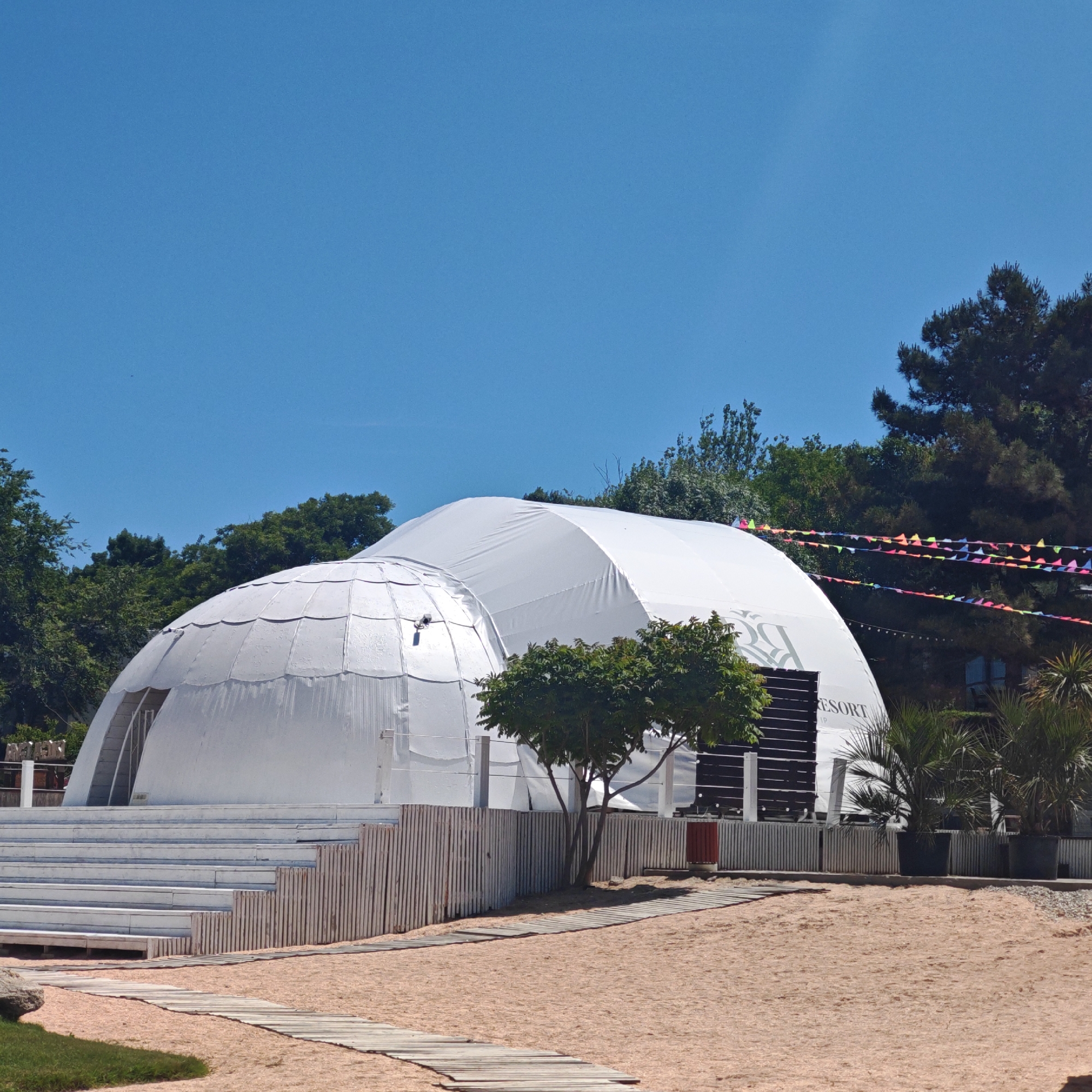 A bright, clear blue sky dominates the upper portion of an outdoor scene featuring a resort or event space. In the center, two large white dome-like structures are prominent. The structure on the left is a smaller, segmented white geodesic dome with an arched entrance, exhibiting what appears to be water streaks or condensation on its surface. Adjacent to it on the right is a larger, smooth white elongated tent structure, displaying a dark logo that reads "RESORT" on its side.
In the foreground, a light-colored sandy area is traversed by a wooden boardwalk pathway, composed of horizontal planks, curving slightly to the right. A small patch of green grass is visible in the bottom-left corner. White tiered wooden steps or seating are positioned in front of the smaller dome. A light-colored wooden slatted fence extends across the middle ground, separating the sandy area from the elevated platform where the structures are located.
Numerous green trees and dense foliage are present, framing the structures from the left, top, and right. A small, multi-trunked tree with green leaves stands between the two white structures, in front of the fence. Several lines of colorful triangular bunting are strung across the upper right section of the image, above the larger tent and trees. On the right side, behind the fence, a red cylindrical bin is visible, along with several potted palm-like plants. No individuals are present in the scene.
Location: Unknown, Unknown.