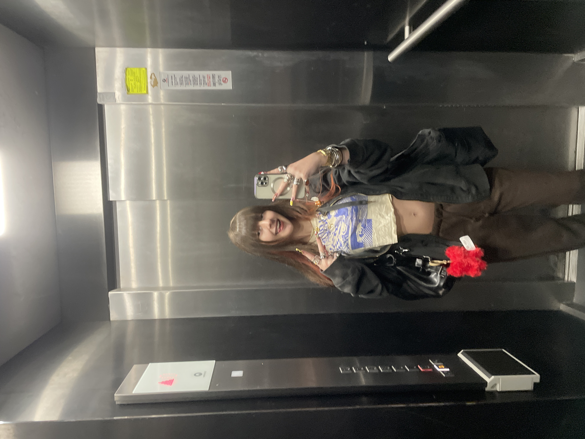 A young woman is taking a selfie in an elevator. She is wearing a black jacket, a light-colored patterned crop top, and brown pants. She is holding a phone in her left hand, which has multiple rings and a bracelet. She has brown hair with red highlights. In the background is the elevator's interior. The elevator has a stainless steel panel with buttons. The location in the city of Raanana, Israel is unknown from the image.
