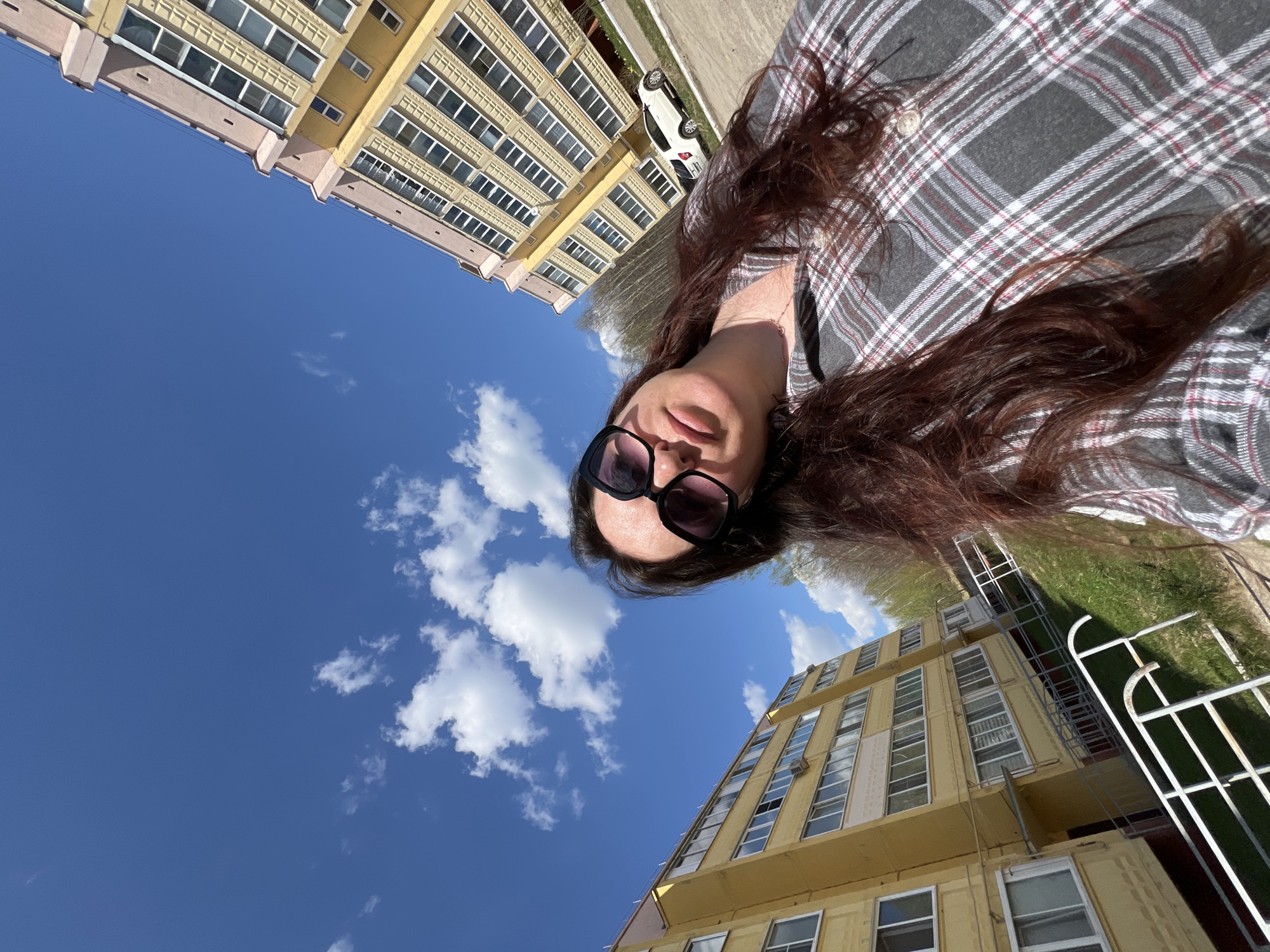 A selfie captures a woman against a backdrop of an urban landscape under a bright, partly cloudy sky. She is wearing sunglasses and a gray plaid shirt, with long, reddish-brown hair cascading around her face and shoulders.
Behind her, the architectural elements dominate the scene. There is a multi-story yellow building with numerous windows, appearing to be a residential or office complex. To the right of the frame, there is a glimpse of greenery, possibly a park or garden, and a white metal railing. A glimpse of a white car is seen on top left of the frame.
The scene is captured on a sunny day, casting light and shadows across the buildings and the sky. The image seems to be taken from a unique angle, looking upward, which skews the perspective of the buildings. Based on these features, it is not possible to determine the location in the city of Noviy, Russia.