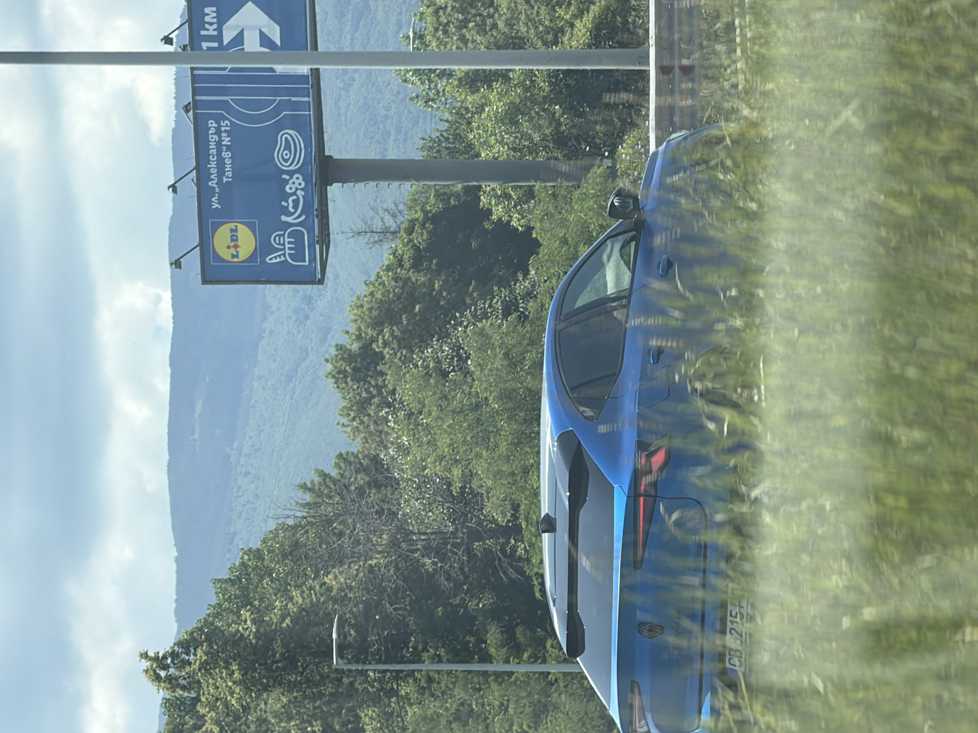 The image shows a blue Renault car driving on a highway with a billboard in the background. The billboard is blue and features the Lidl logo, along with text that appears to be in Bulgarian: "ул.,, Александър Танев № 15" and "1 км" with a directional arrow. The car's license plate is visible and reads "CB 3215 FO". The environment is green with trees and hills in the distance, under a cloudy sky. The scene is likely located in or near Sofia, Bulgaria, given the Bulgarian text on the billboard and the typical highway scenery associated with the city's outskirts.