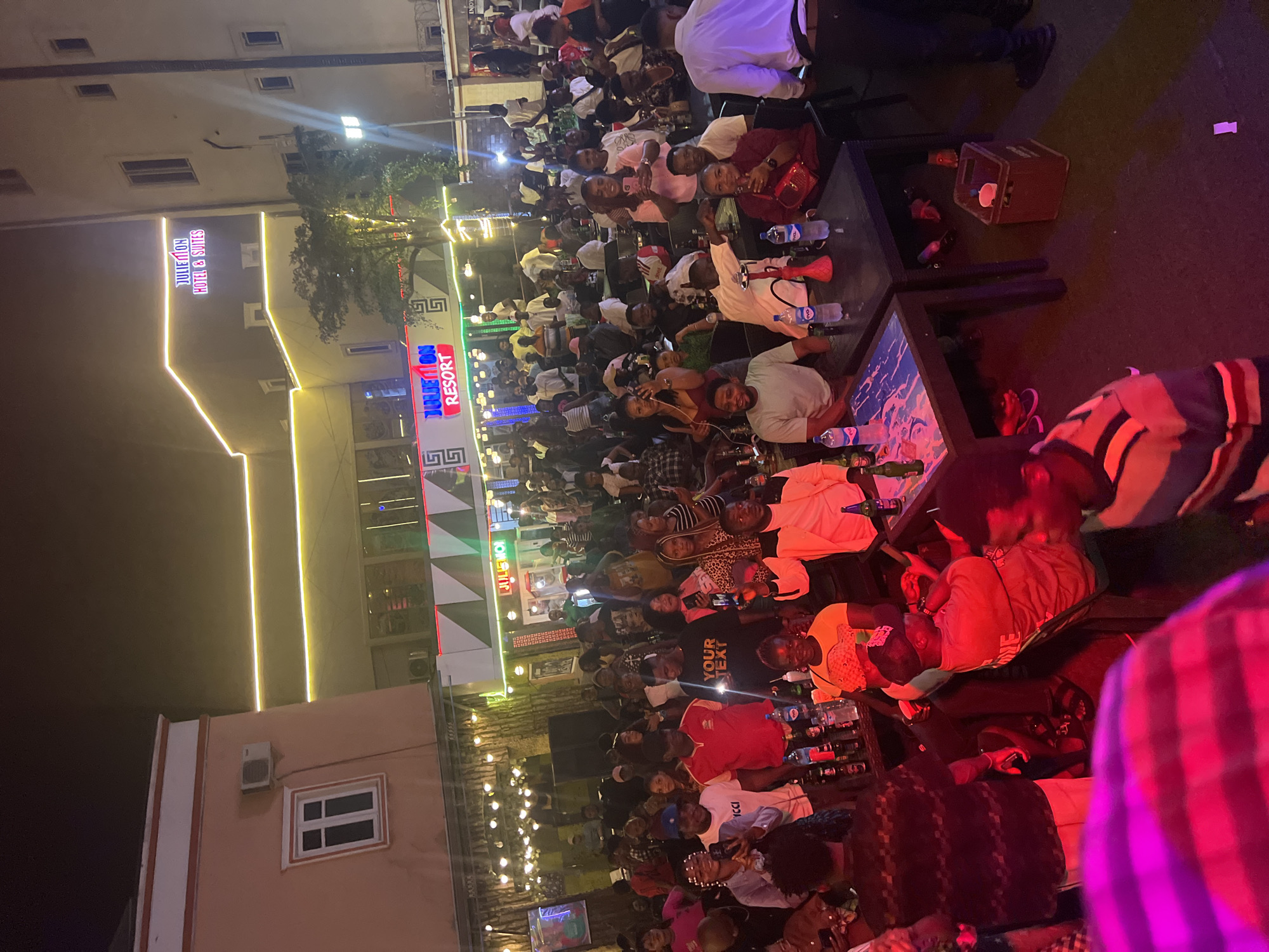 The image depicts a lively outdoor gathering at Juleimon Resort in Oba, Nigeria. A large crowd of people, predominantly young adults, are socializing, some standing and others seated at tables. The atmosphere is festive, indicated by the presence of drinks on tables and the general buzz of activity. The resort features prominent signage with the name "Juleimon Resort" illuminated by neon lights. The architecture includes multi-story buildings with windows, and decorative lighting strands are strung across the outdoor area, suggesting a nighttime or evening event. Many individuals are holding drinks or phones, and some are engaged in conversation. The foreground is partially obscured by a blurred section of what appears to be a person in brightly colored clothing under intense pink lighting. The ground is carpeted, and various tables and seating arrangements are visible throughout the scene.