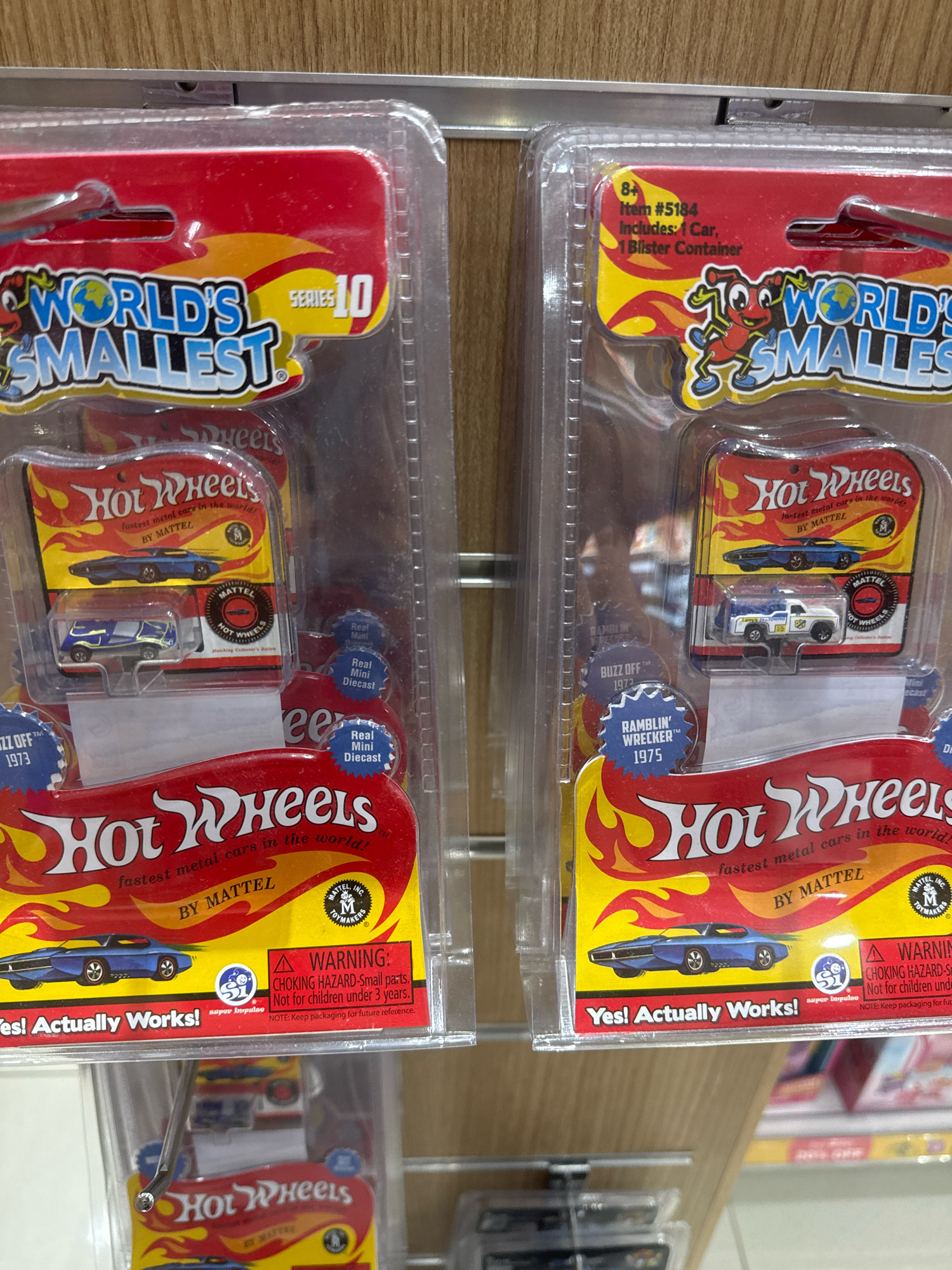 The image displays two blister packs of Hot Wheels "World's Smallest" series 10 toy cars, arranged side-by-side on a retail shelf. Each blister pack features a red backing card with "World's Smallest" text in a stylized font, a graphic of a racing car, and a smaller blister containing a miniature diecast car. The pack on the left contains a blue car, and the pack on the right contains a white and blue tow truck. Text on the packaging includes "Hot Wheels fastest metal cars in the world!" and "by MATTEL". Warning labels related to choking hazards are also visible. The background shows wooden shelving and other retail items, suggesting a store environment. The image does not provide any information about the city of Al Matar, United Arab Emirates, or any specific location within it.