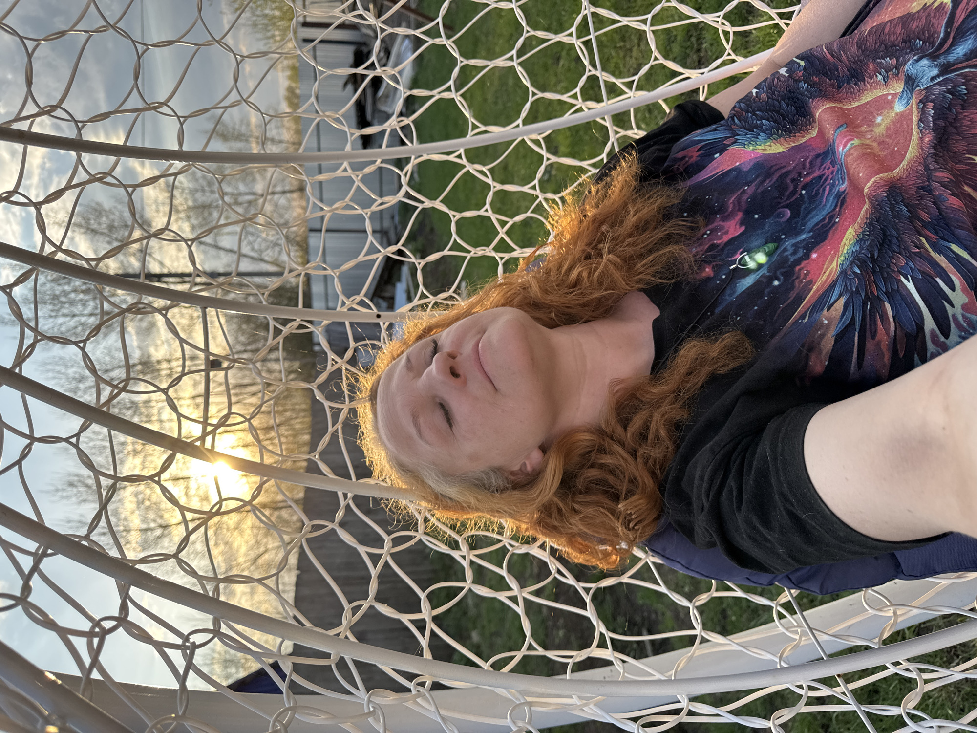 A woman with light skin and reddish hair is lying in a white, net-like hanging chair. She is wearing a black shirt with a colorful graphic of a phoenix. Her eyes are closed, and her face is relaxed. The sun is setting in the background, visible through the netting of the chair. The location may be Chaikovsky, Russia, but this cannot be confirmed from the image. Other background elements include trees and a building.