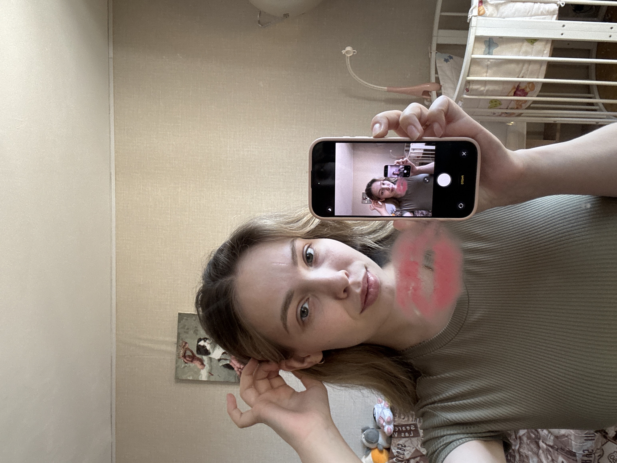 A young woman is holding a smartphone and taking a photo of herself in a bedroom. The woman has fair skin, light brown hair, and is wearing a ribbed olive-green shirt. The phone in her hand displays a mirrored image of the same scene. The background includes a light beige wall, a crib with bedding, a small picture hanging on the wall, and what appears to be a cluttered floor. It's difficult to determine the location is specifically Chaikovsky, Russia, from this image alone.