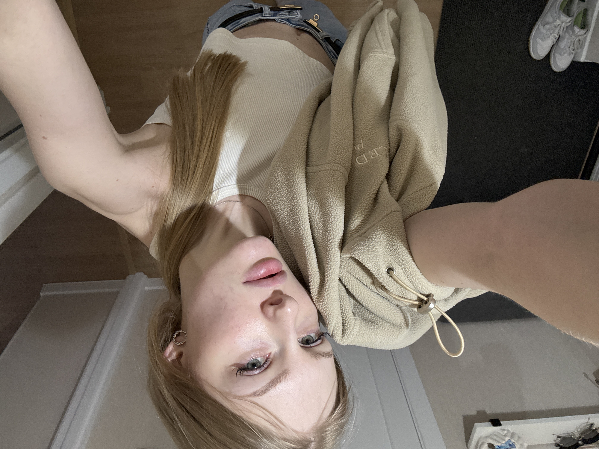 A self-portrait captured from an overhead angle depicts a young woman looking directly at the camera. Her head is positioned towards the bottom of the frame, with her long, light brown hair fanning out around her face. She has fair skin, light-colored eyes (appearing blue or green), and full, lightly tinted lips. She wears a circular, possibly jeweled, earring in her visible ear.
Her attire consists of a white ribbed tank top and blue jeans, fastened with a black belt that has a gold rectangular buckle. A beige or tan fleece jacket or sweatshirt is draped over her, partially covering her torso. Faint text reading "CED po" (likely part of a brand name or word) is visible on the fleece, which also features a drawstring with a silver toggle.
The setting is indoors, appearing to be a casual room in Perm, Russia. The background includes a light brown wooden floor, a dark mat or rug in the upper right, and what looks like a white wall or door. A pair of white sneakers with subtle green accents rests on the dark mat. In the lower right corner, a surface or shelf is partially visible, holding small items such as sunglasses. The image primarily captures a casual pose with no specific activity or event other than the act of taking a picture.