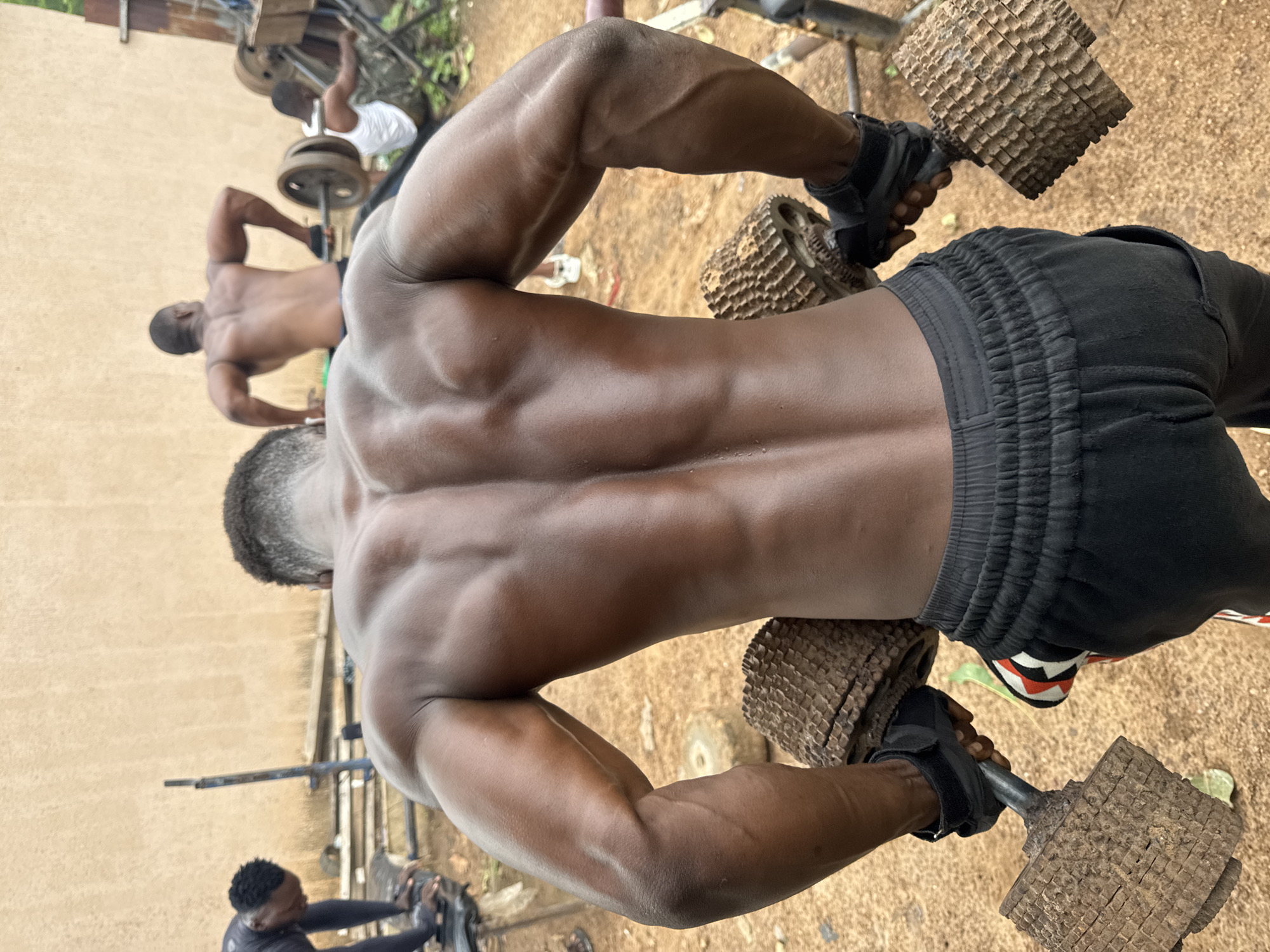In Akwanga, Nigeria, a man with dark skin and a muscular physique is seen from behind, performing a weightlifting exercise outdoors. He is wearing black shorts with a red and white trim on the side of one leg. His hands, clad in black weightlifting gloves, grip two heavy, rustic-looking dumbbells. These dumbbells appear to be constructed from wood and metal, with a textured, weathered surface. The man's back muscles are prominently defined, showcasing his strength.
In the background, other individuals are also engaged in exercise. To the left, another man, also with dark skin, is bent over a weight bench, wearing a black long-sleeved shirt. Further back and to the right, a third man, wearing a white tank top, is lifting a barbell. The environment appears to be a makeshift outdoor gym, with dirt ground and rudimentary weightlifting equipment visible. A plain, textured wall forms the backdrop. The overall scene depicts a moment of intense physical training in a communal, outdoor setting.