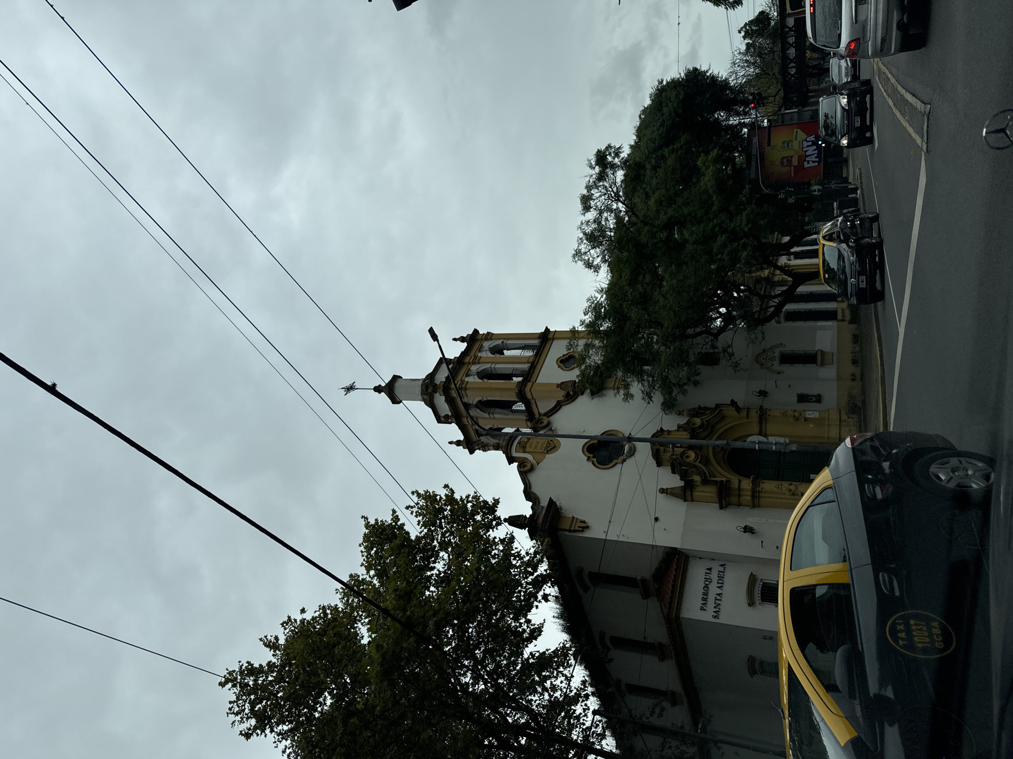 The image shows a street view in Buenos Aires, Argentina, featuring the Parroquia Santa Adela church. The church has a white and yellow facade and a visible bell tower. Overhead power lines stretch across the sky, which is overcast. A black and yellow taxi is prominent in the foreground, partially obscuring the lower right portion of the church. Several other vehicles are visible further down the street, along with trees lining the road. A billboard with the word "Fanta" on it is positioned across the street.