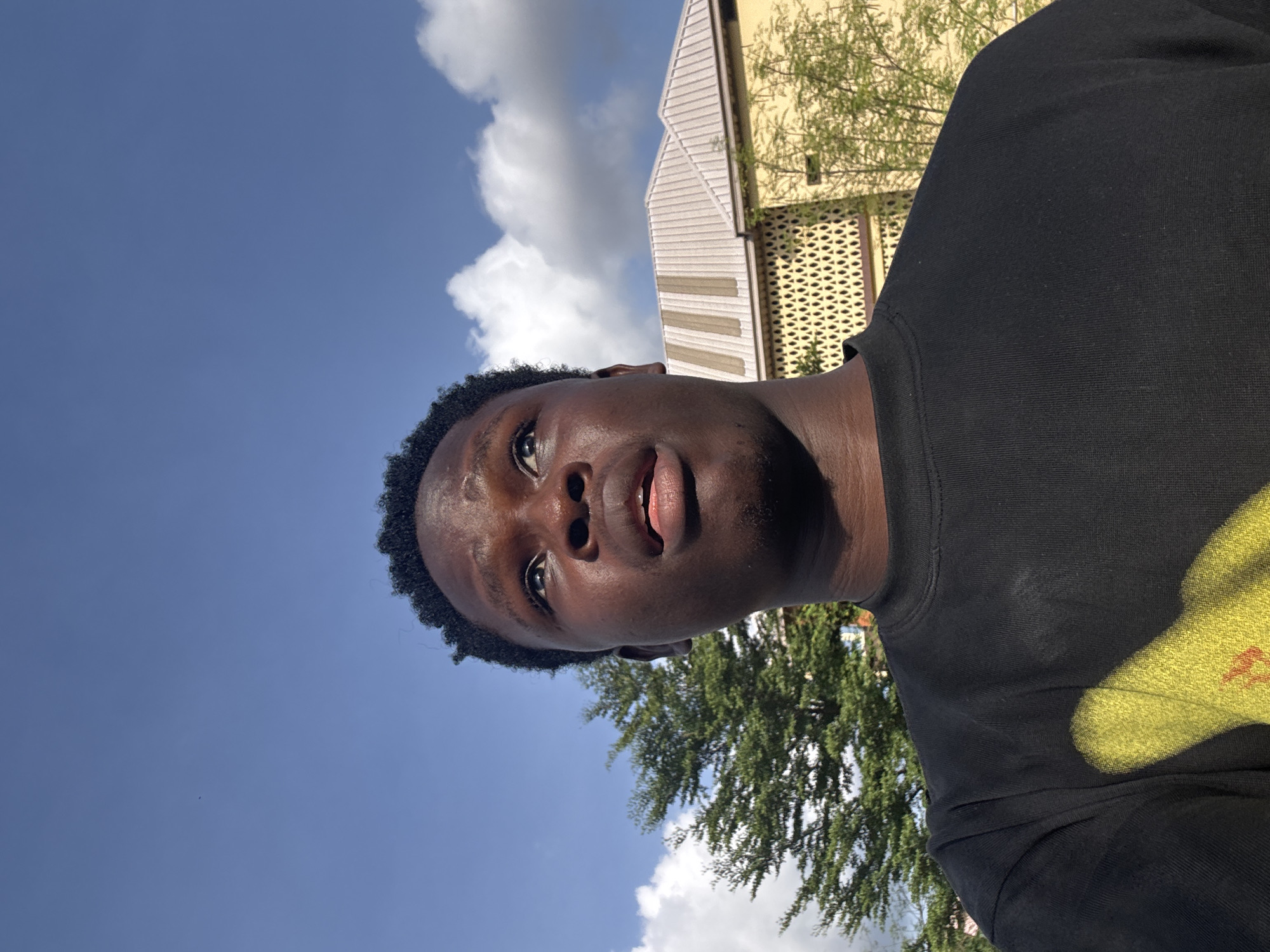 A young Black male is captured in a close-up, low-angle selfie, looking upwards towards a bright blue sky with scattered white clouds. He is wearing a black t-shirt with a faded yellow graphic design on the left side of his chest. Behind him, to the left, is the lush green foliage of a tree, and to the right, a portion of a building with a light-colored, pitched roof and yellow walls featuring a distinctive patterned facade. The scene suggests an outdoor setting in Enugu, Nigeria.