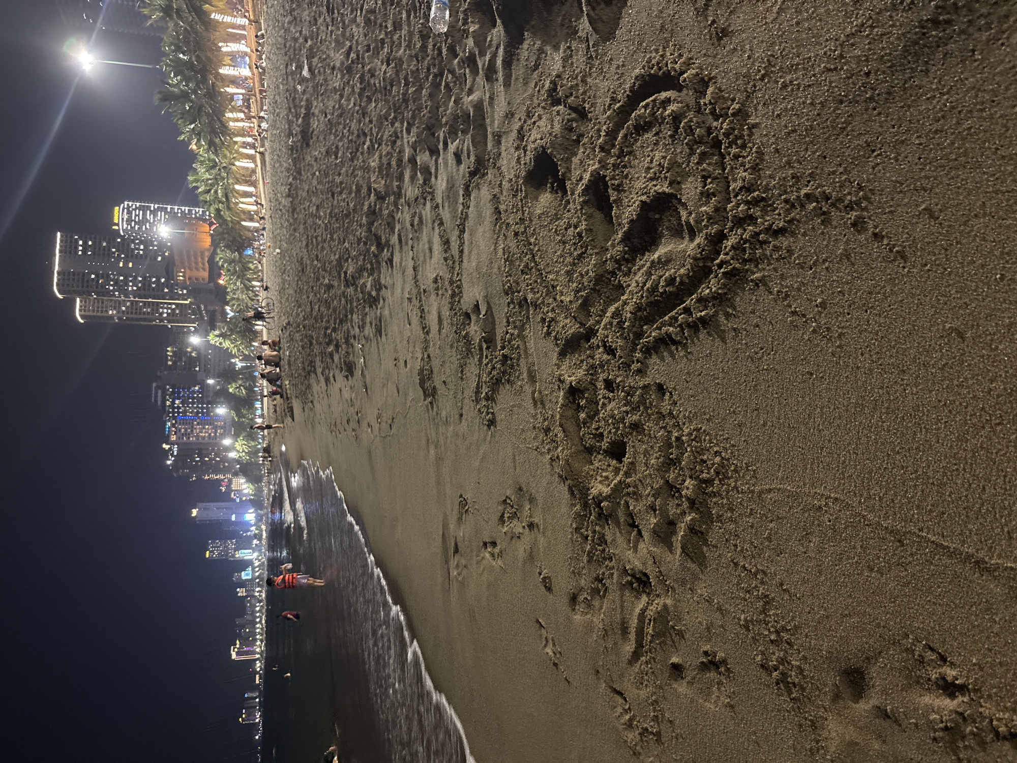 At night, a sandy beach in Nha Trang, Vietnam, features the Nha Trang coastline illuminated by numerous lights from high-rise buildings and palm trees adorned with decorative lights. The ocean is dark, with gentle waves lapping the shore, and a foamy white line marks the water's edge. Several individuals are in the shallow water and on the wet sand, some wearing orange life vests. On the dry sand in the foreground, a drawing of a smiley face is visible. Further down the beach, more people are scattered across the sand, and a plastic bottle lies on the sand. The sky is dark and clear.